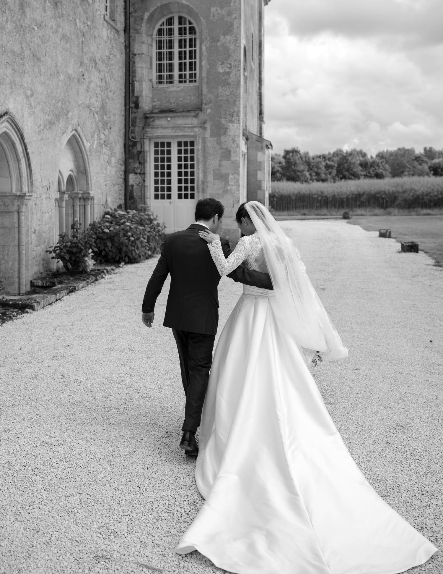 Elegant couple walks along historic stone building in black and white.