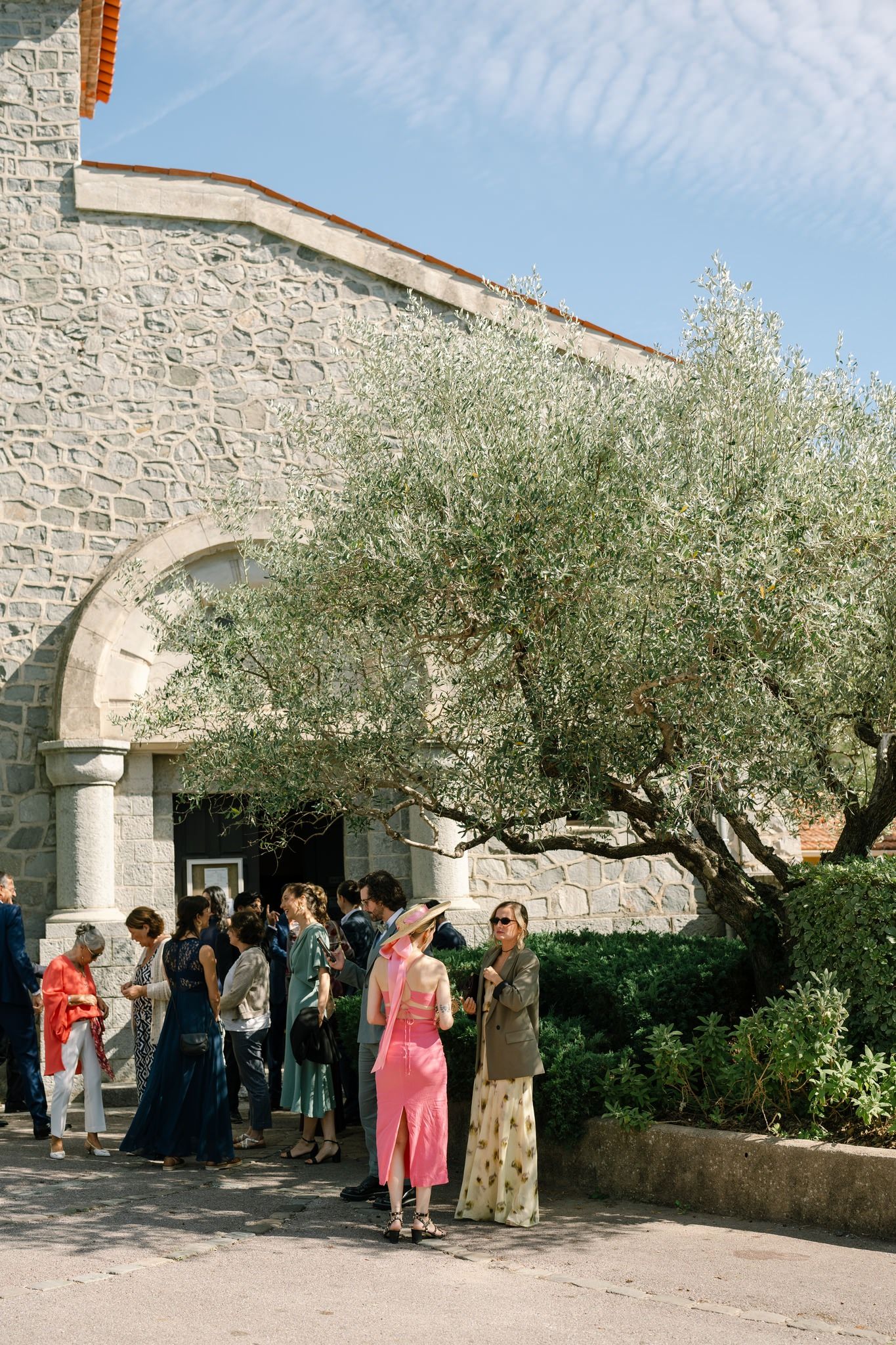 Wedding guests gathering outside a rustic stone church under olive trees.