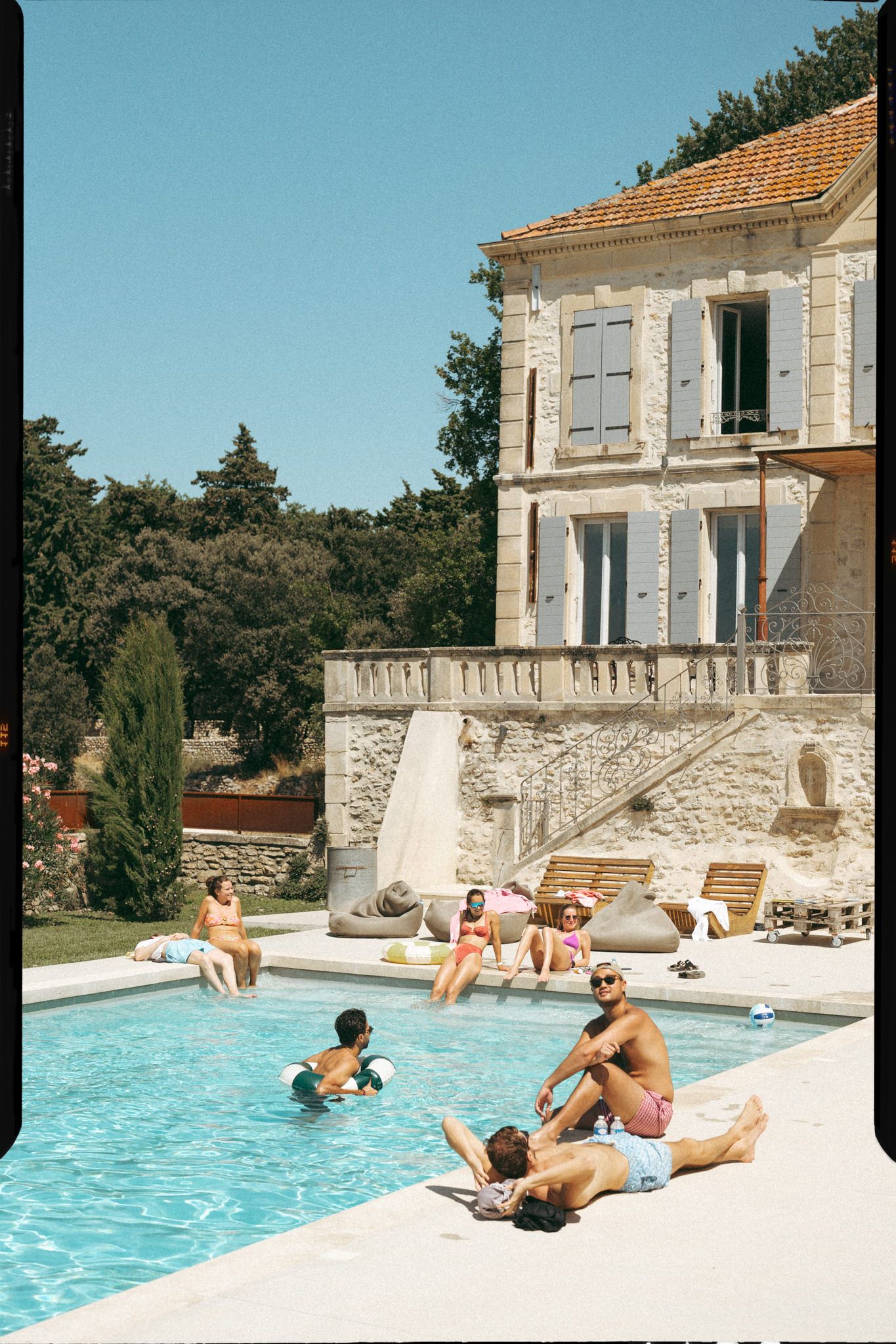 People lounging and swimming by a luxurious stone villa pool on a sunny summer day in Mediterranean setting.