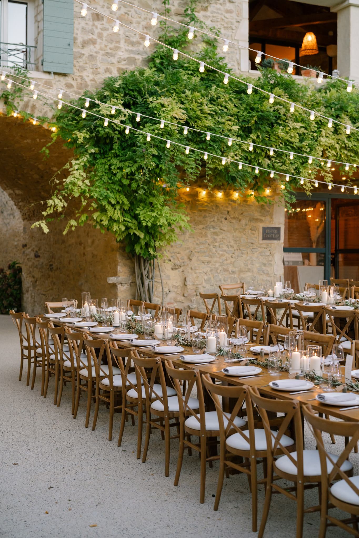 Rustic outdoor wedding reception setup with wooden chairs and string lights at a stone venue.