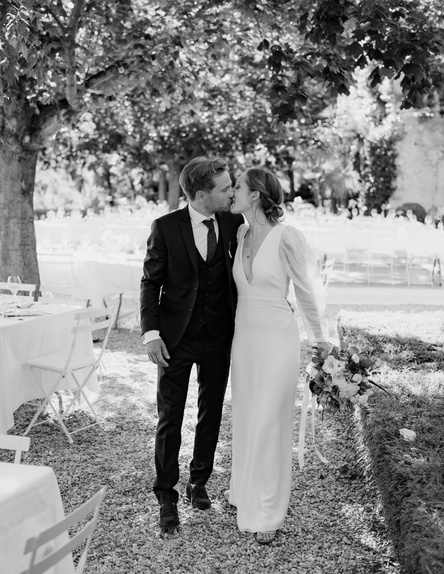 Black and white wedding portrait showing a couple walking through a garden with white chairs visible.
