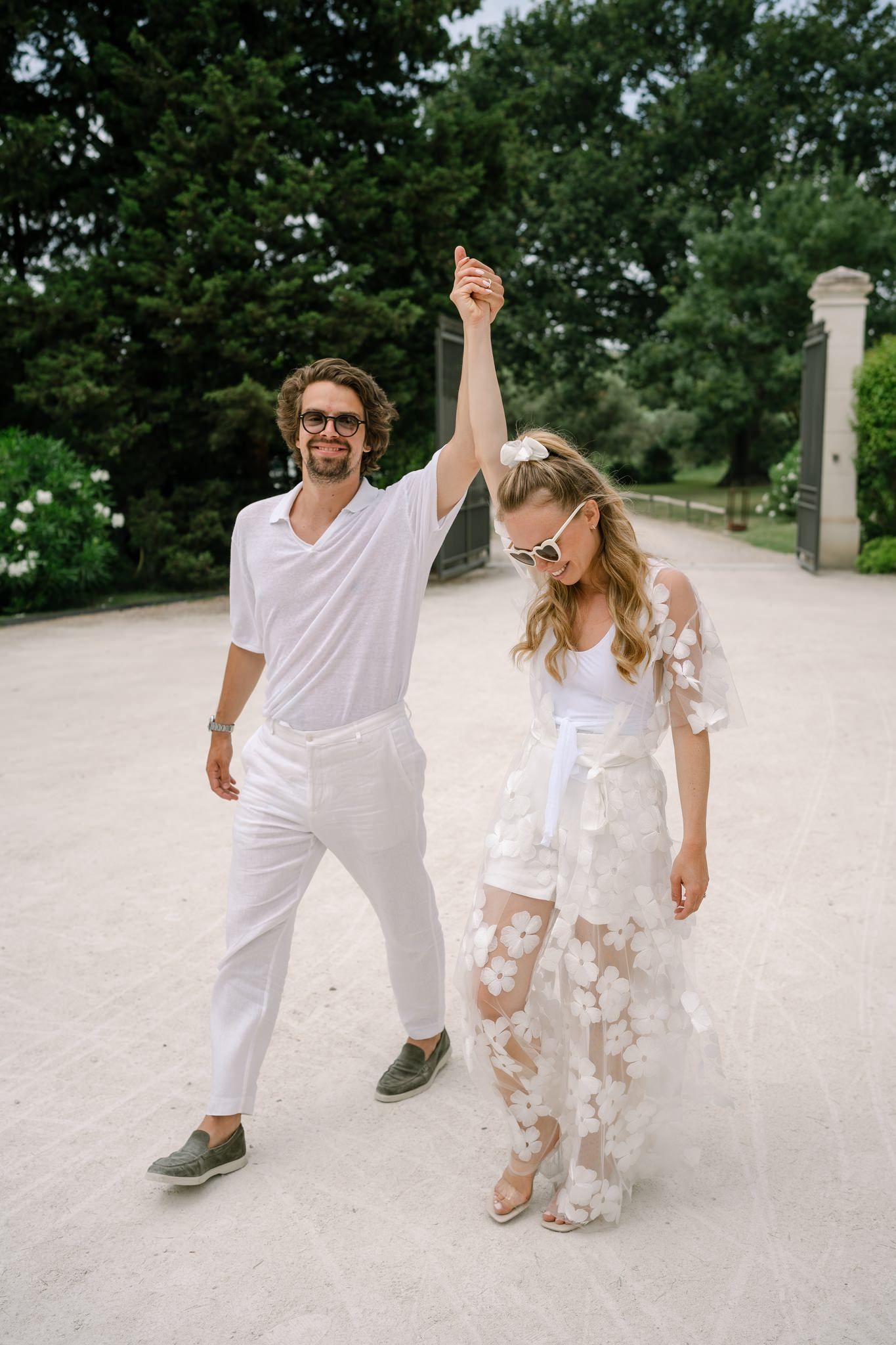 Playful couple dancing on white pathway surrounded by green trees.
