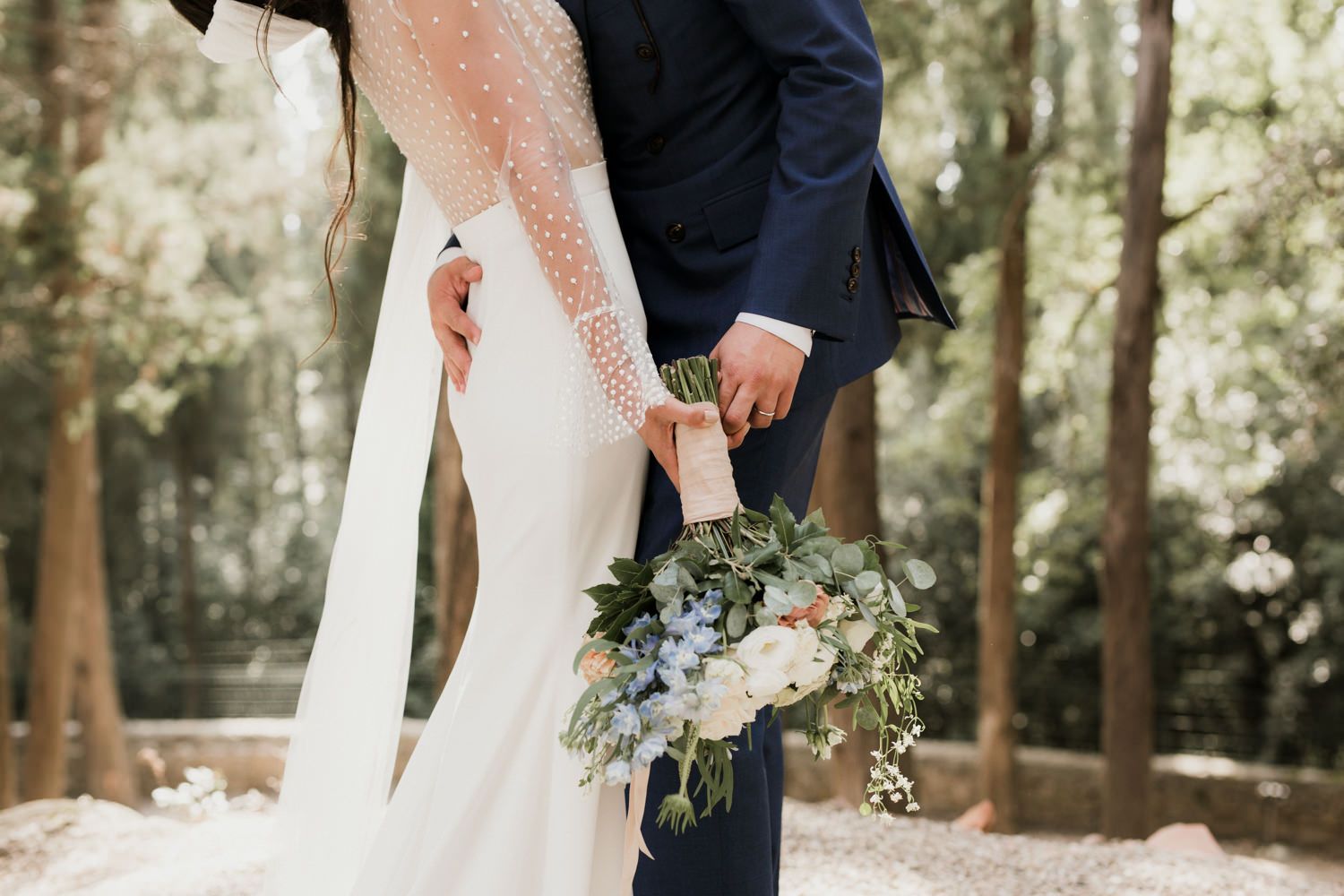 Bride holding a delicate bouquet poses with groom among towering trees.