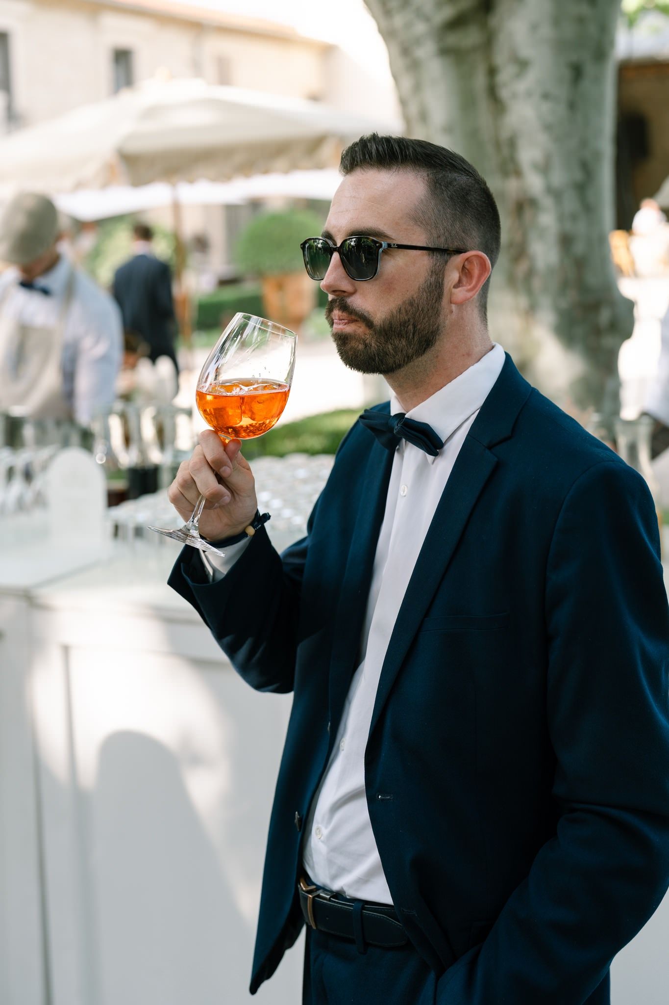 Stylish guest in a dark suit and sunglasses enjoying a cocktail at an outdoor reception.