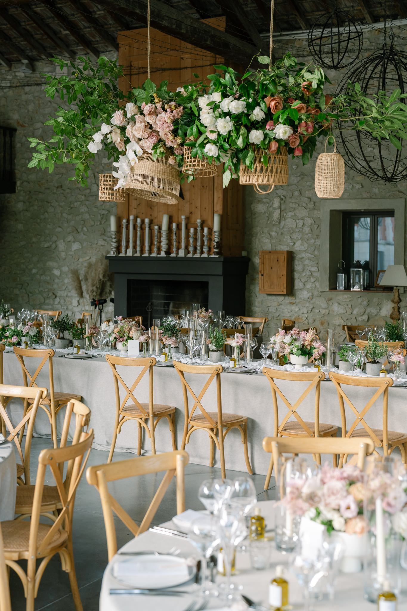 Elegant wedding reception table setting with hanging floral arrangements and wooden cross-back chairs in a rustic venue.