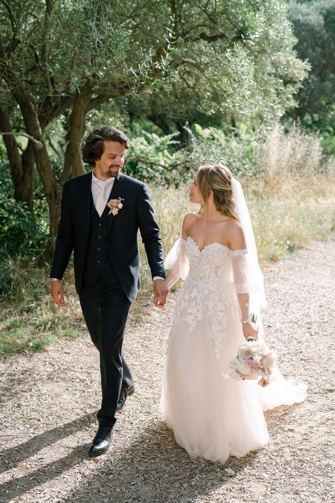 Newlyweds walking together on rustic gravel path surrounded by olive trees.