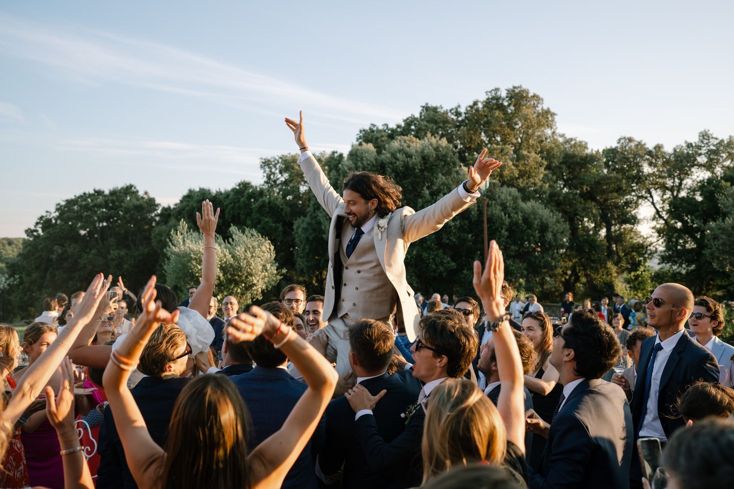 Wedding couple lifted in celebration by cheering guests during outdoor reception.
