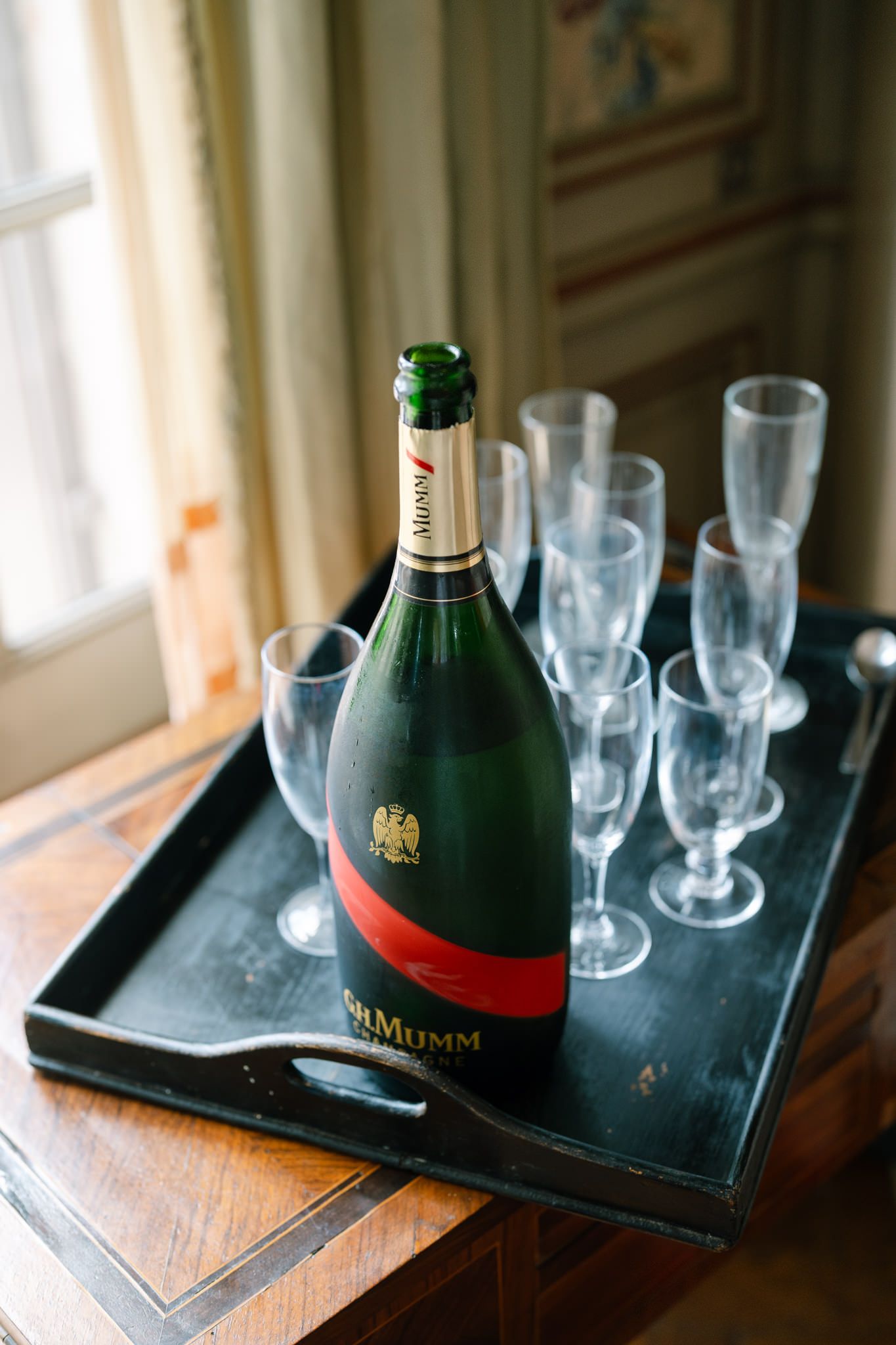 Champagne bottle and crystal glasses arranged on dark serving tray near window.