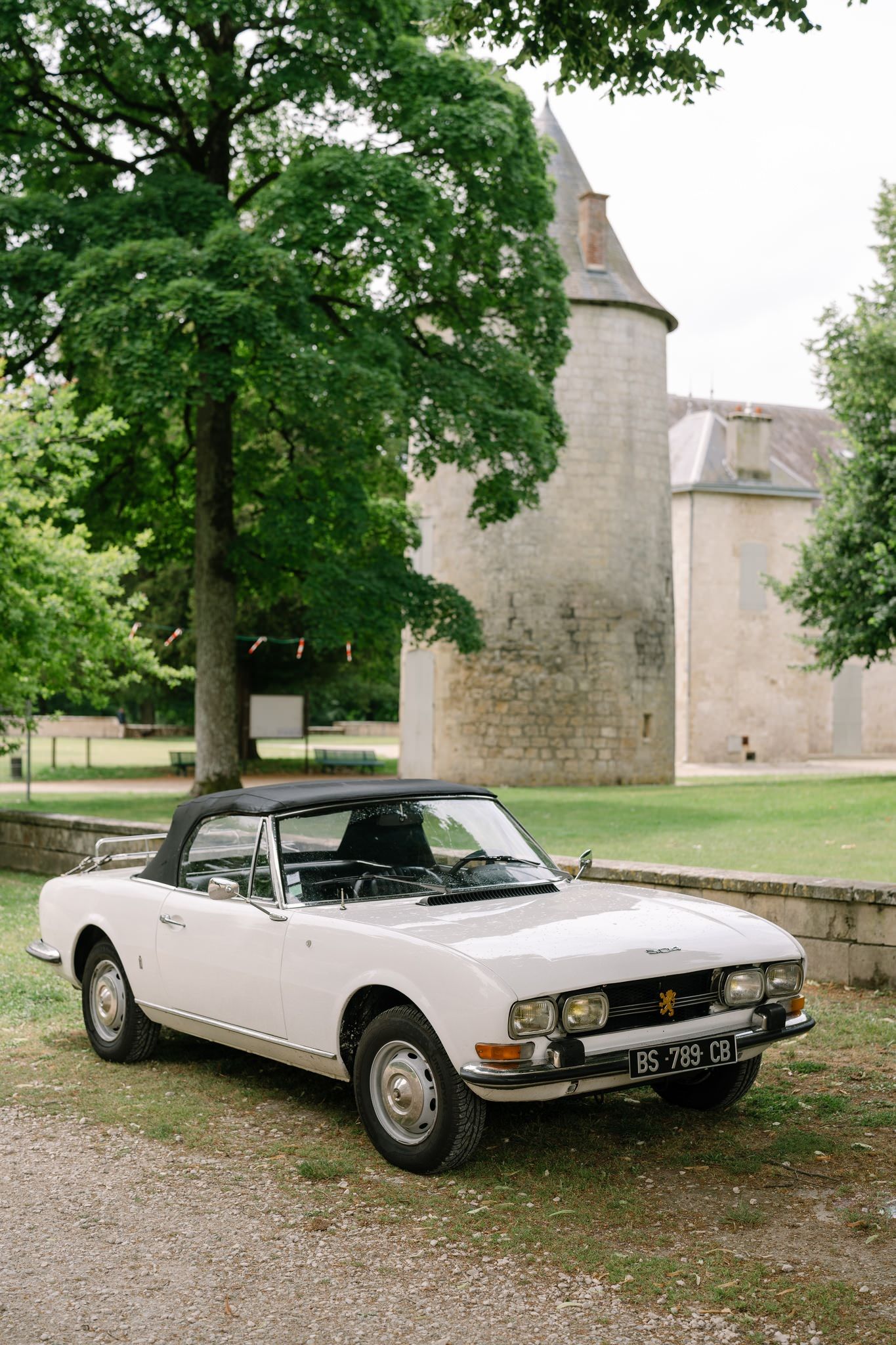 Classic white convertible car parked outside a stone building under green trees.