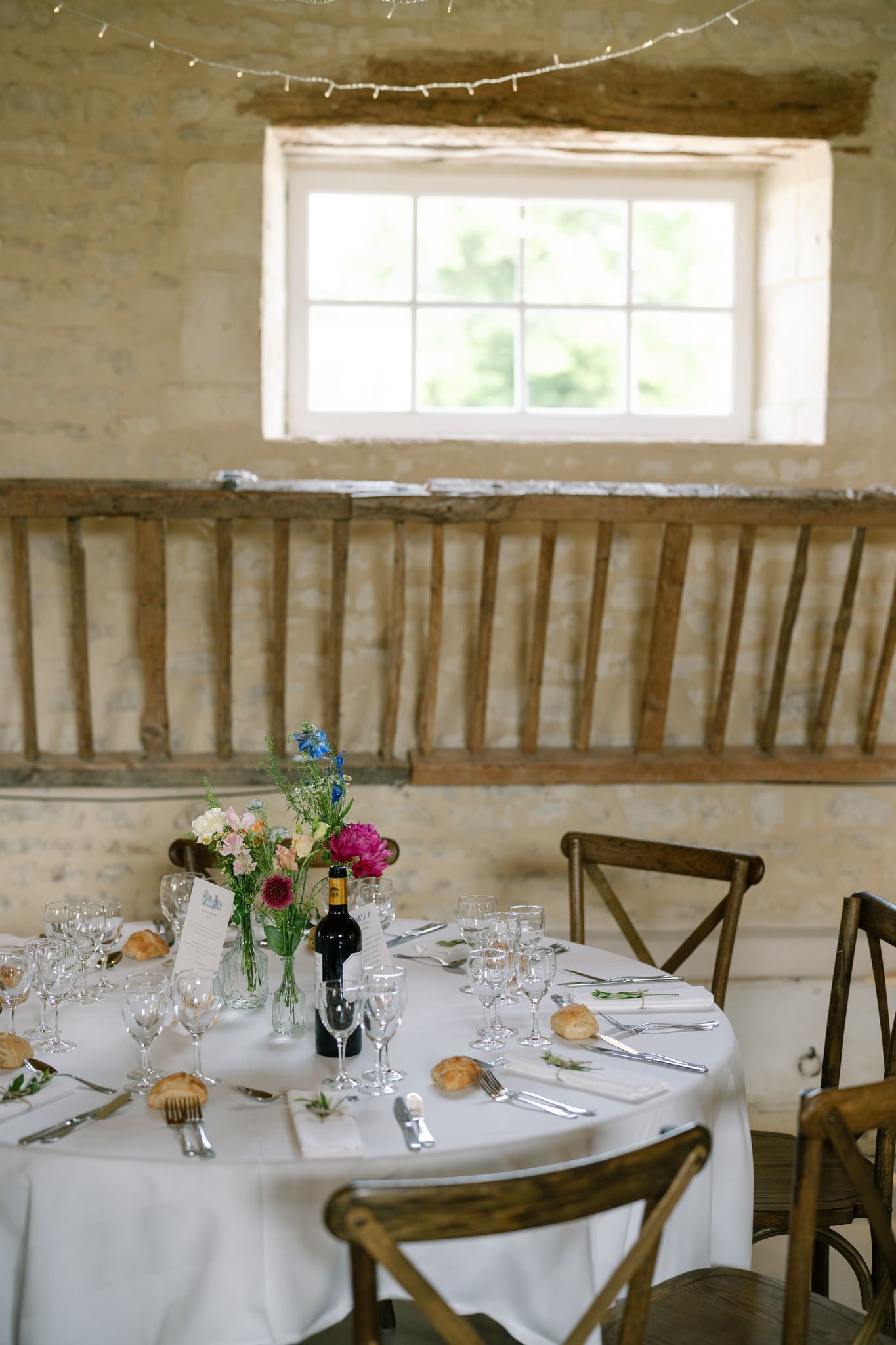 Rustic wedding reception room with stone walls, white tablecloths and wooden cross-back chairs.