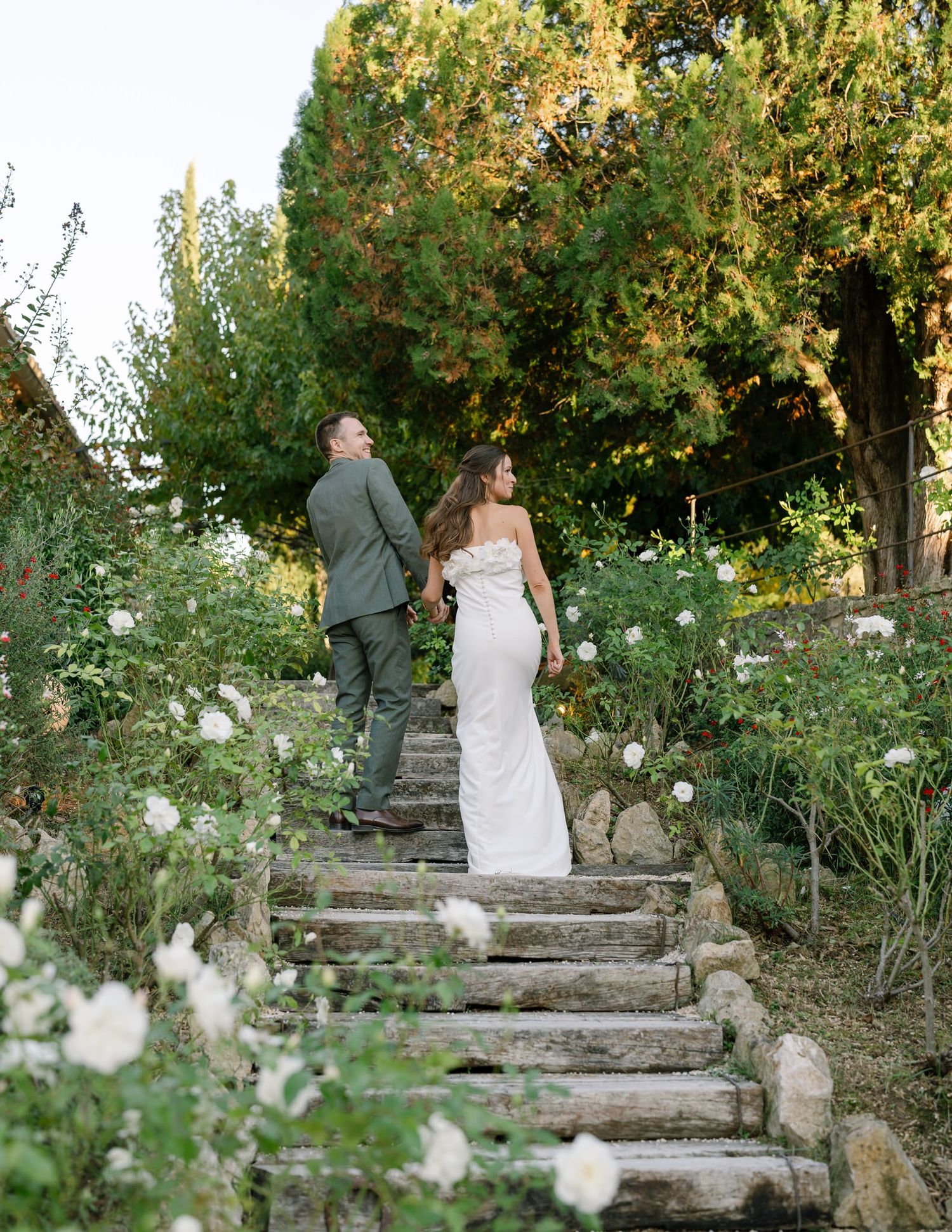 A couple ascends stone garden steps surrounded by white flowers and greenery.