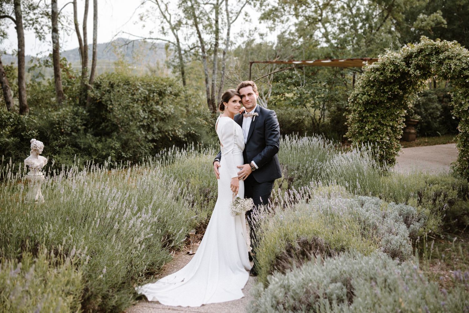A wedding couple poses in a lush garden setting with lavender plants and greenery.