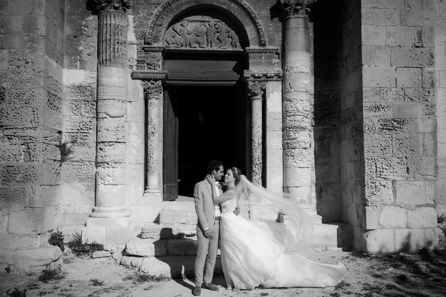 A romantic wedding photo in black and white featuring a grand stone archway entrance with ornate columns.
