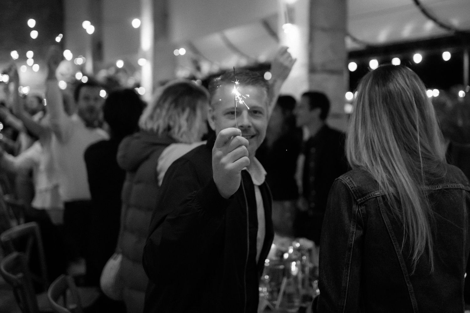 Wedding guests dance and celebrate on a dimly lit dance floor with string lights overhead.