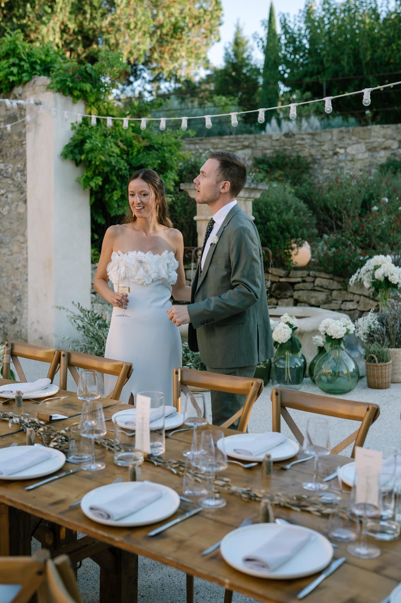 Rustic outdoor wedding reception table setup with string lights and wooden chairs.