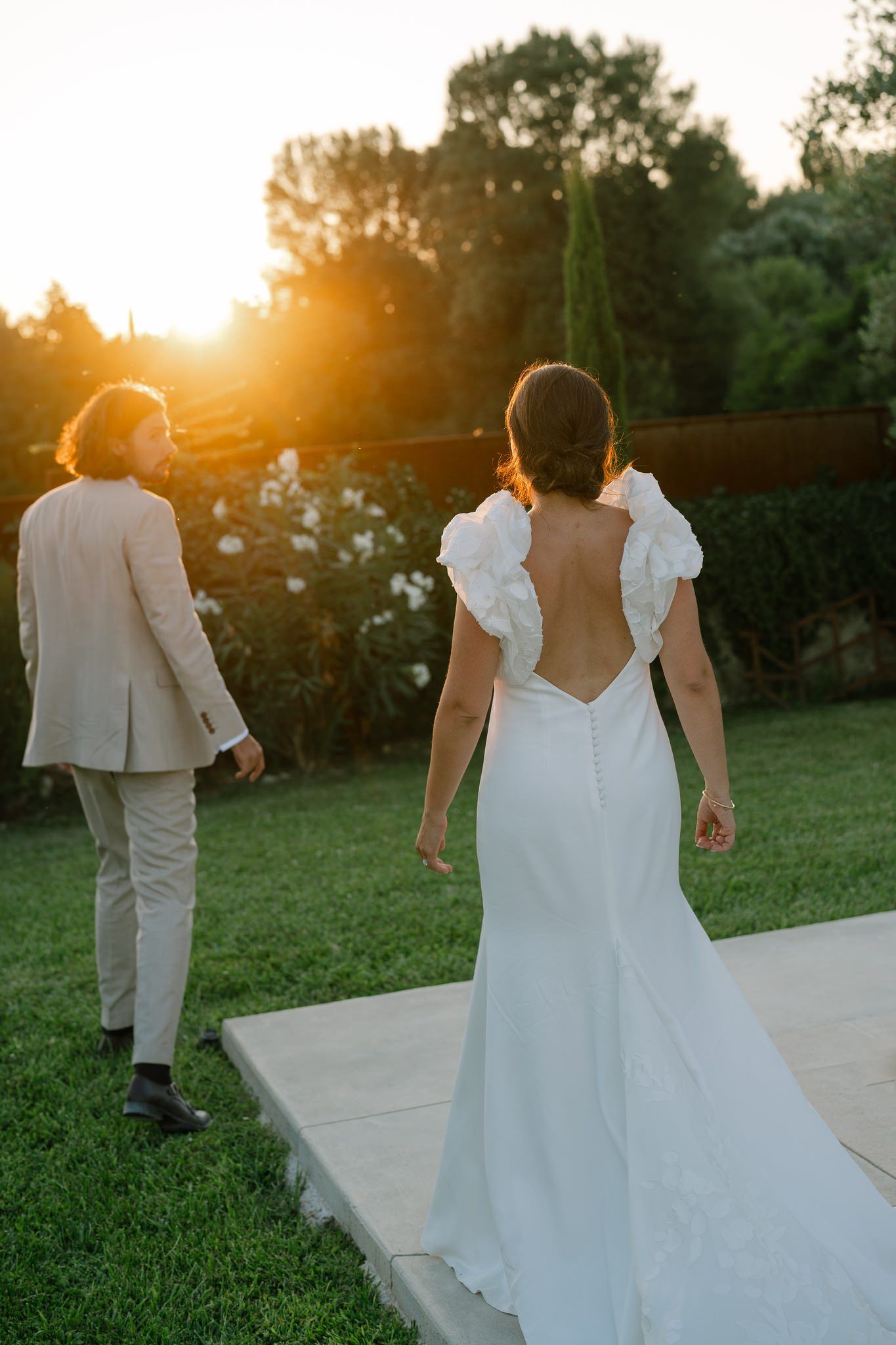 Elegant wedding dress with open back design silhouetted against sunset in garden setting.