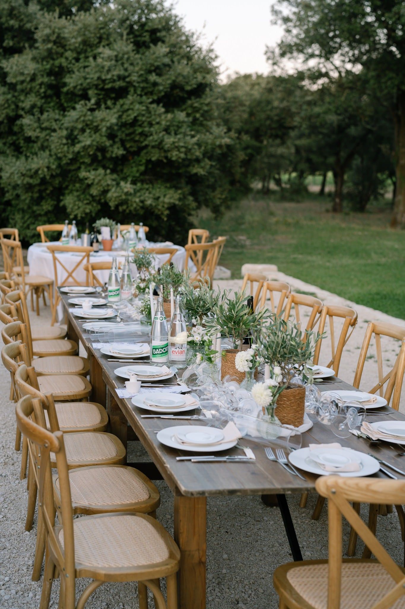 Long wooden dining table with crossback chairs set for an outdoor wedding reception.