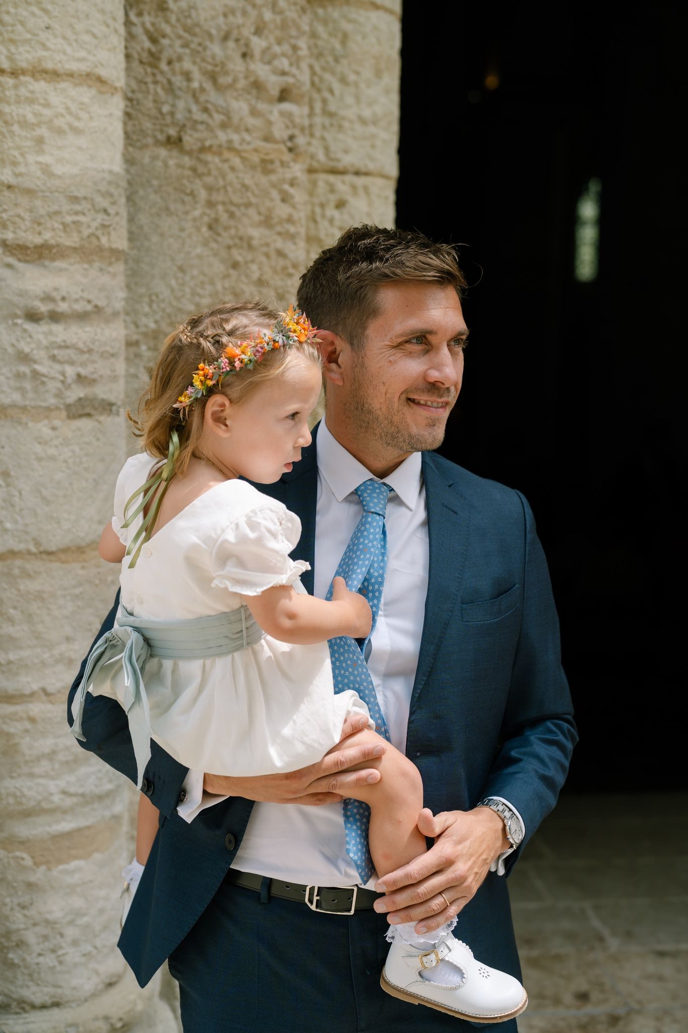 Sweet moment between wedding guest and young child in white dress outside stone church.