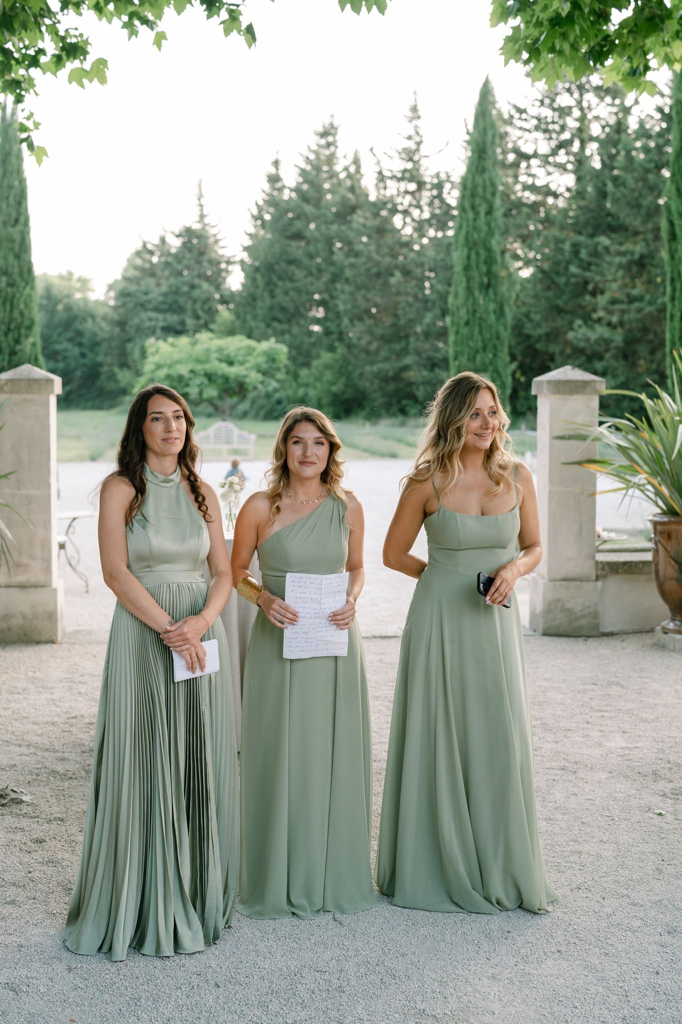 Three bridesmaids in sage green floor-length dresses standing outdoors.