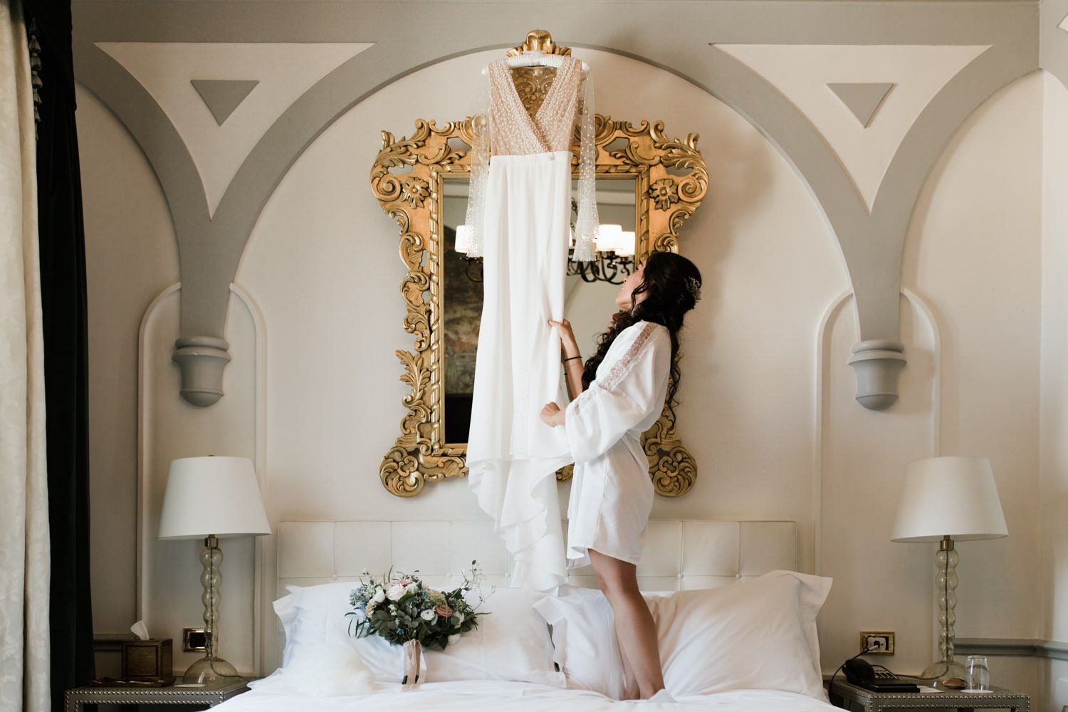 A wedding dress hangs from an ornate gold mirror in an elegant bedroom setting.