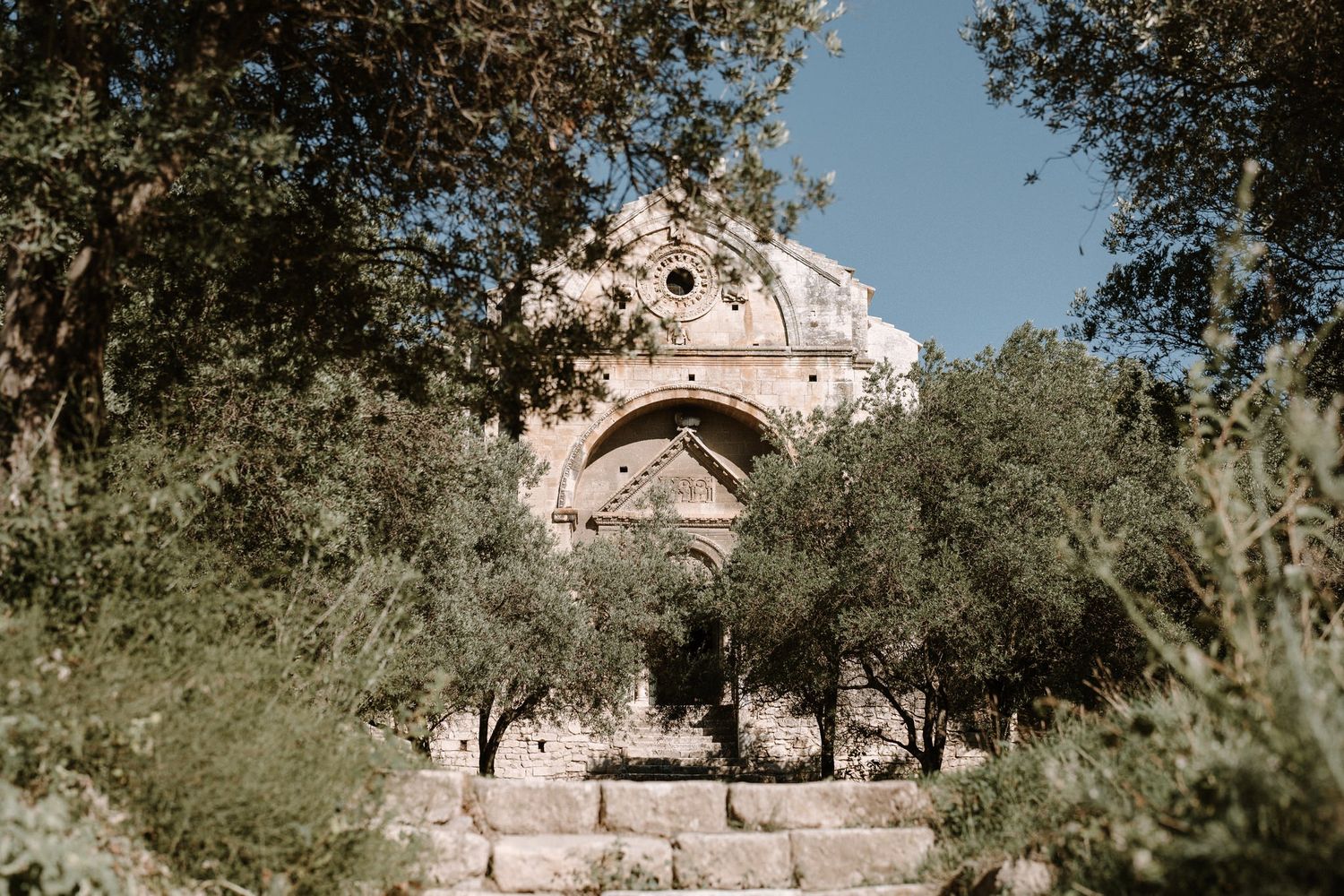 Ancient stone church with arched architecture surrounded by olive trees under blue sky.