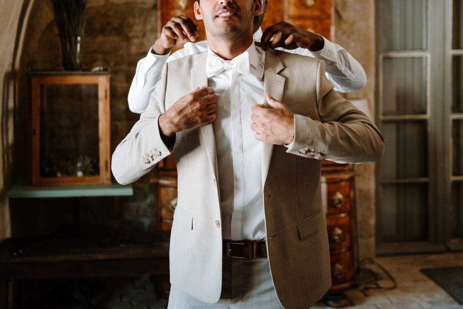 A groom adjusts his tie while getting ready in a rustic indoor setting with warm lighting.