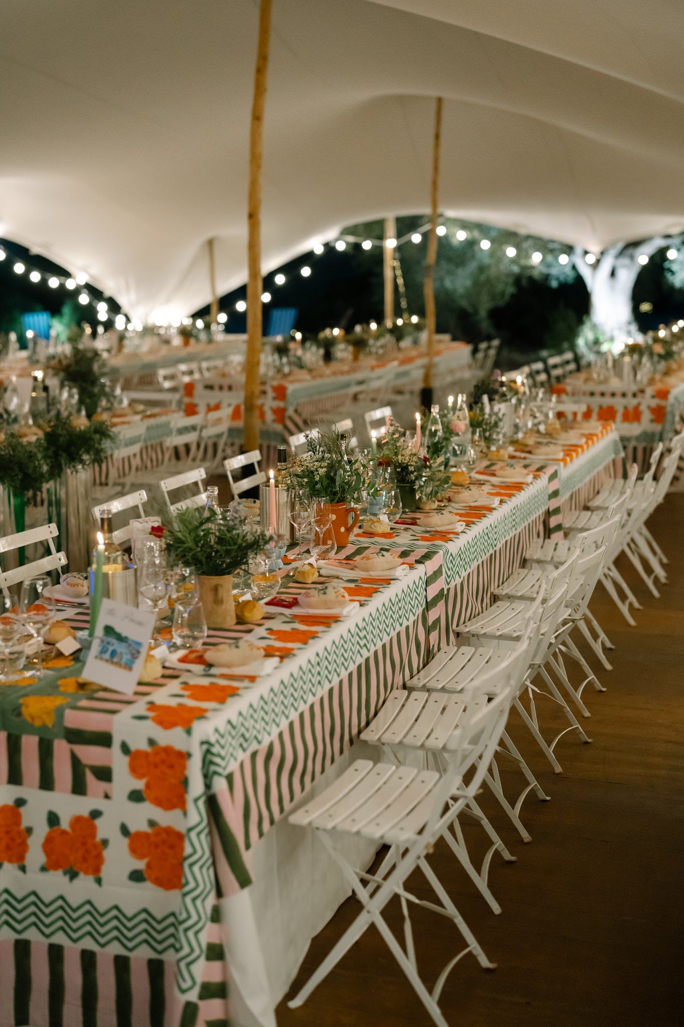 Long reception tables set with white chairs and orange accents under a white tent.
