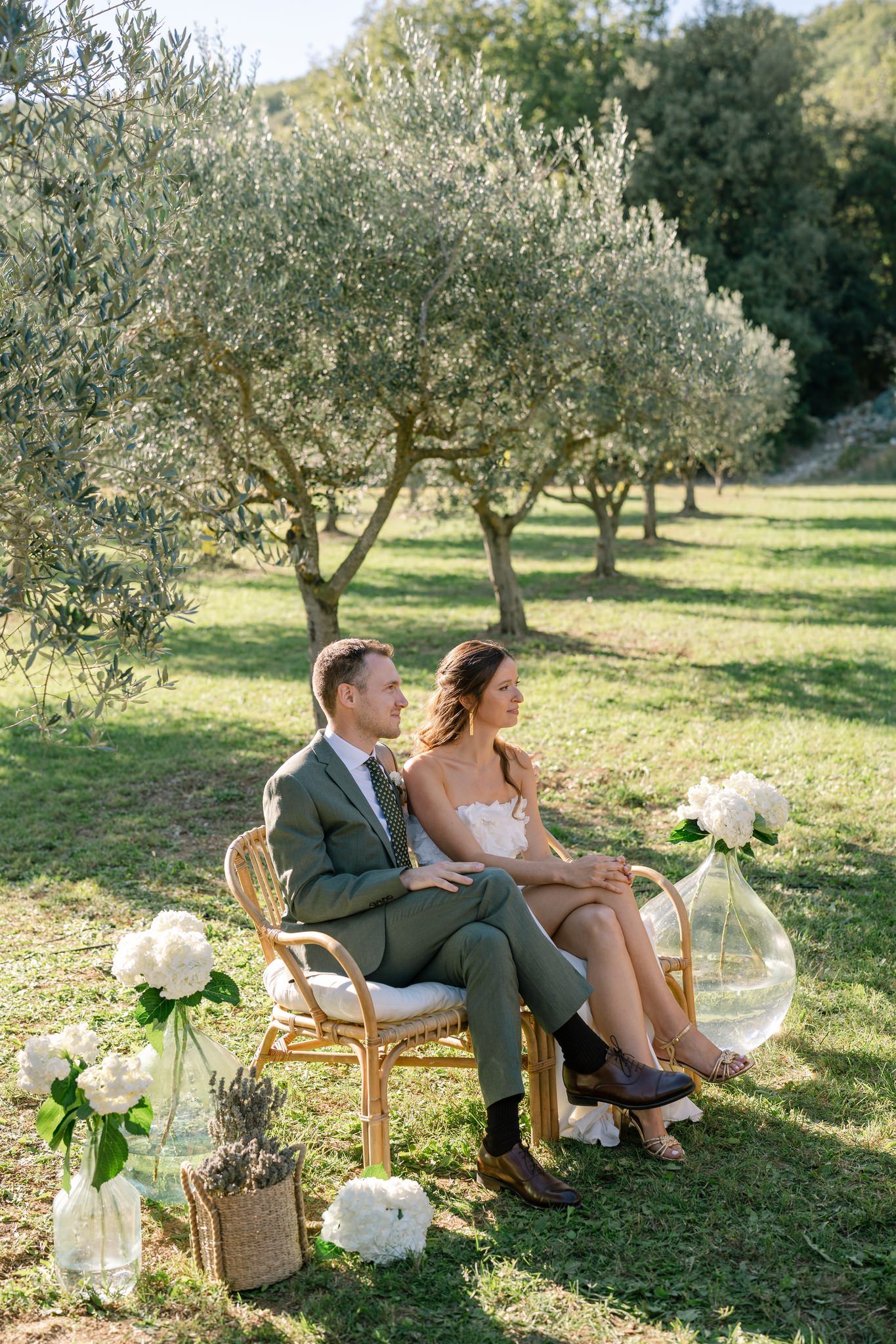 A couple relaxes on vintage chairs surrounded by floral arrangements in an olive grove.
