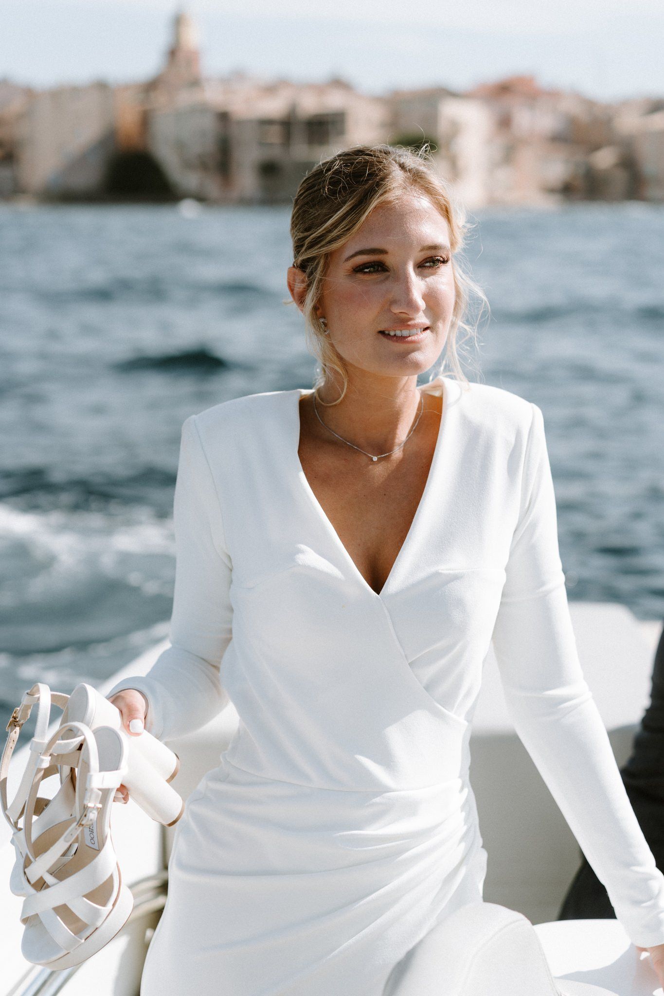 Woman in elegant white long-sleeved wedding dress poses by waterfront.