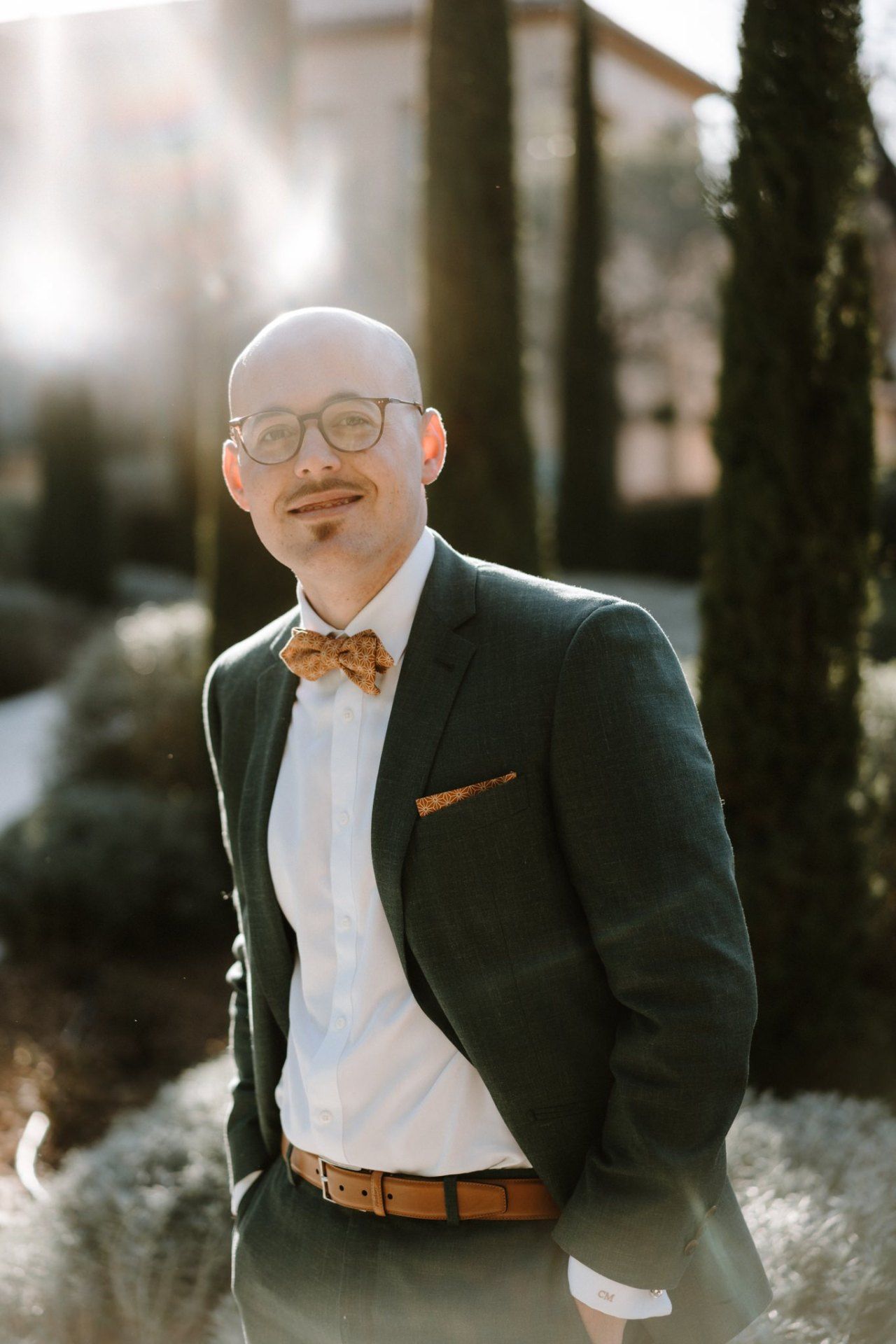 A formal portrait in a black tuxedo with bow tie and brown leather belt against a natural backdrop.