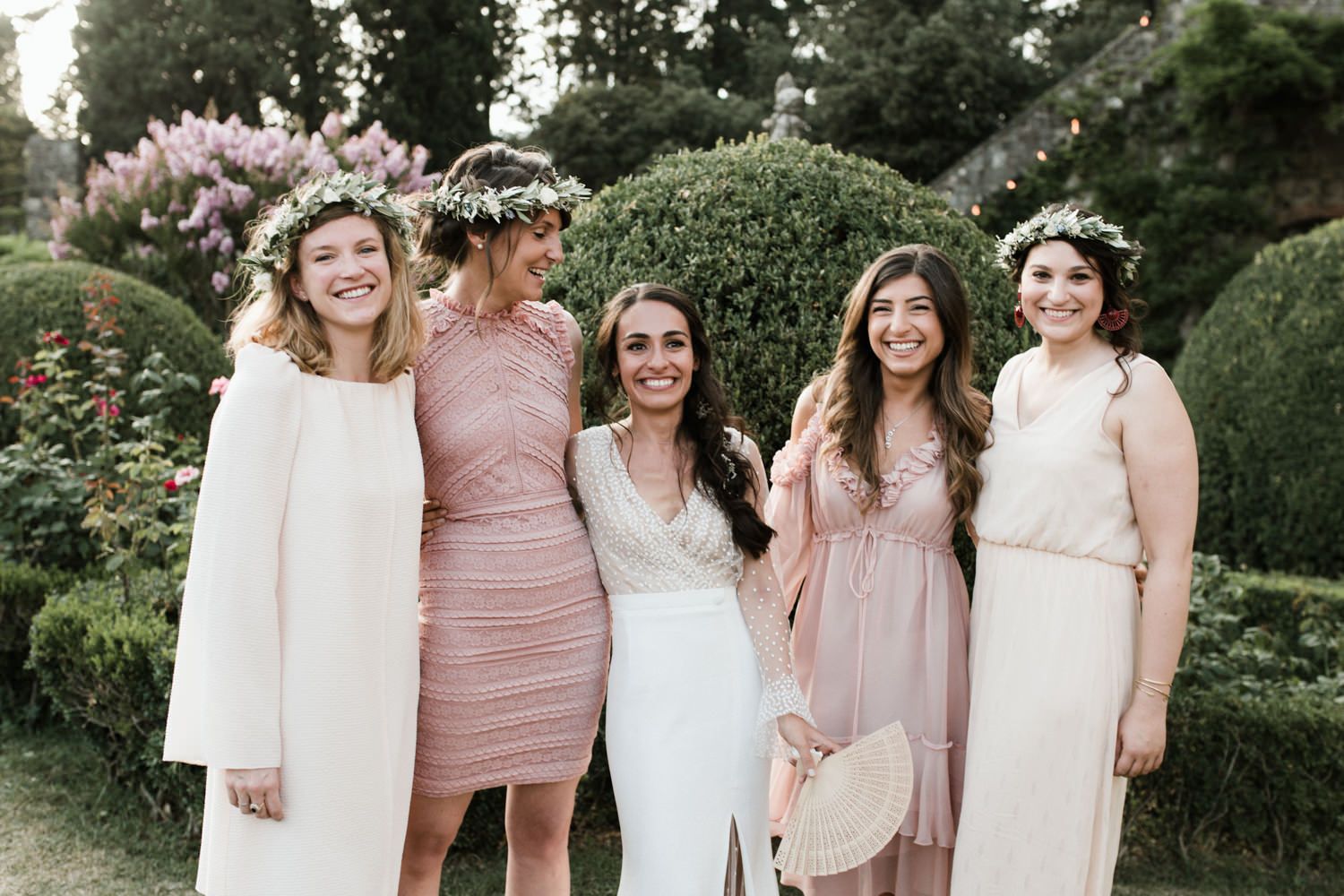 Wedding party wearing blush and cream colored dresses poses together in a garden setting.