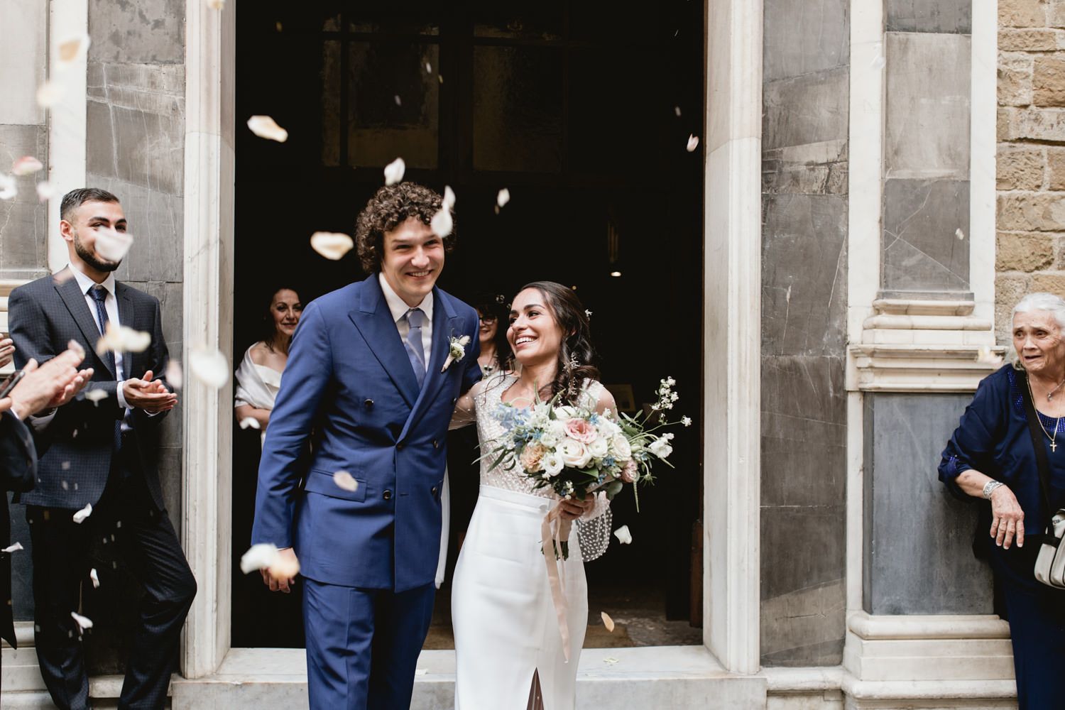 Newlyweds exit church while guests throw flower petals in celebration.