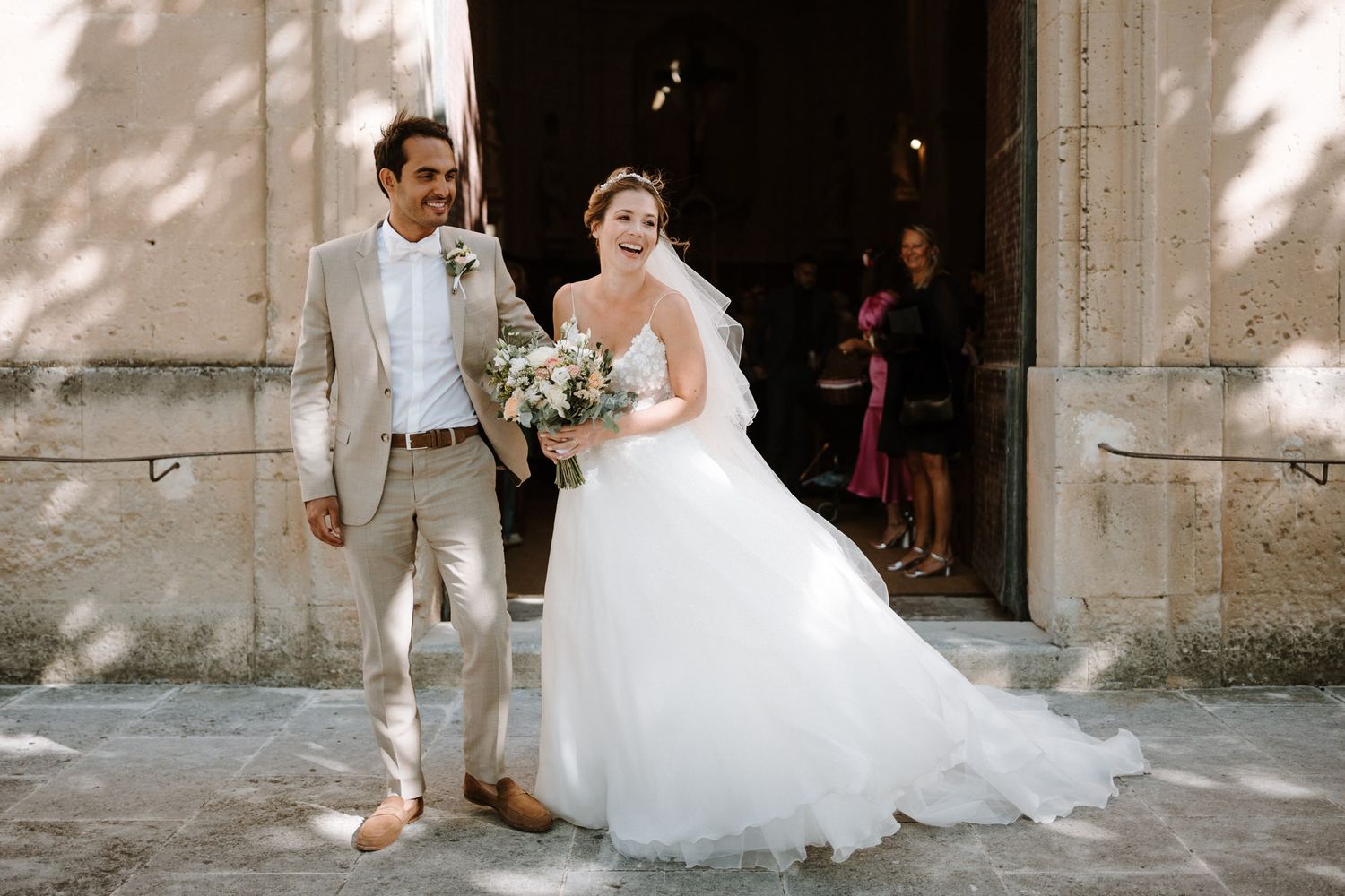 A wedding couple exits a stone church on a sunny afternoon.