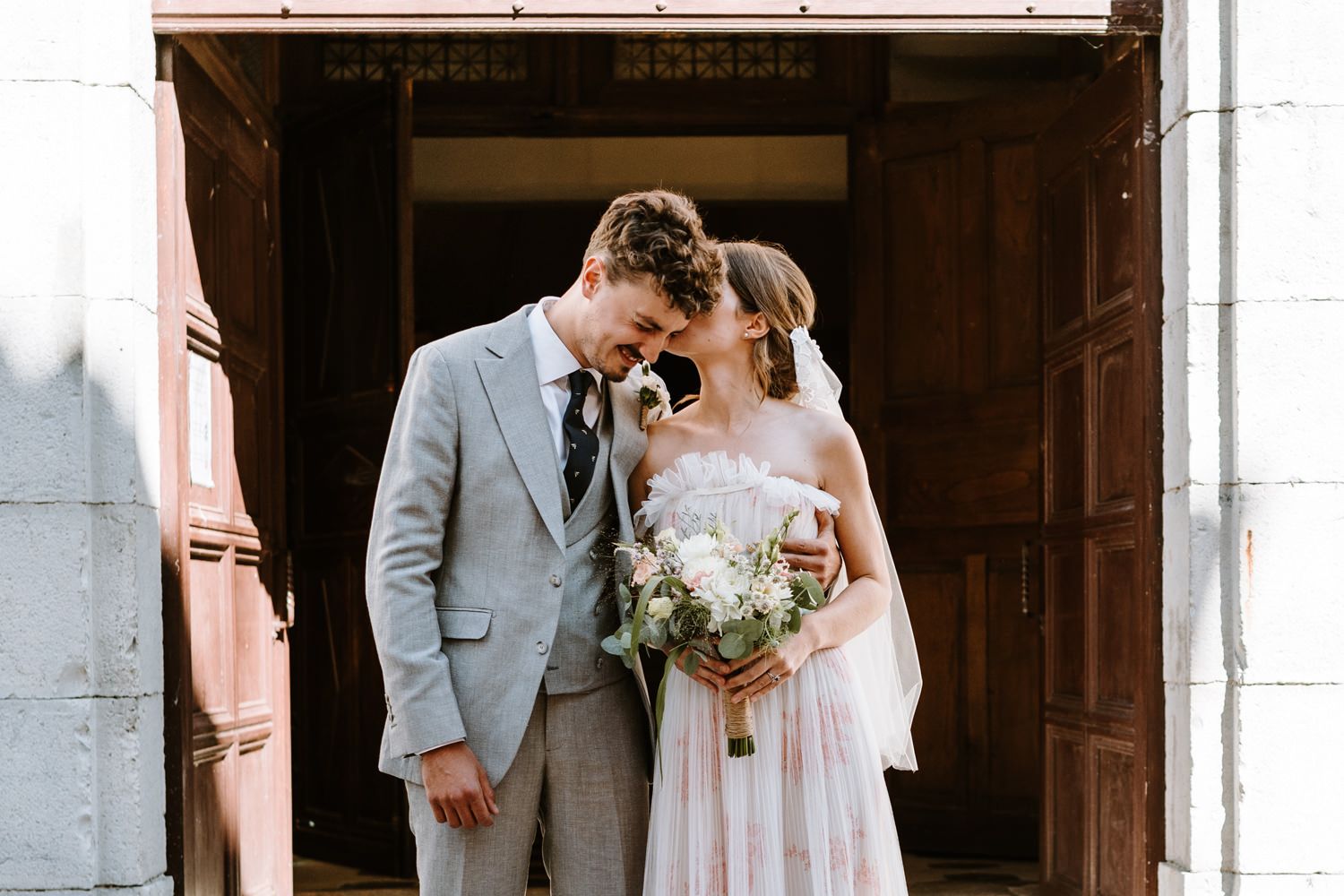 A romantic wedding portrait of a couple with bouquet in front of historic church doors.