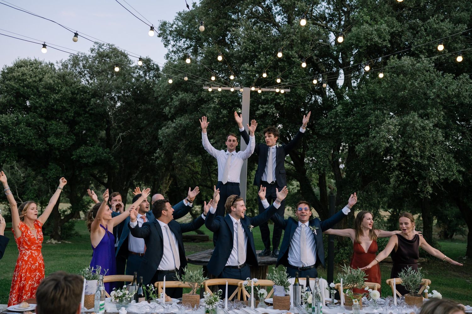 Wedding guests raise their hands in celebration under string lights at dusk.