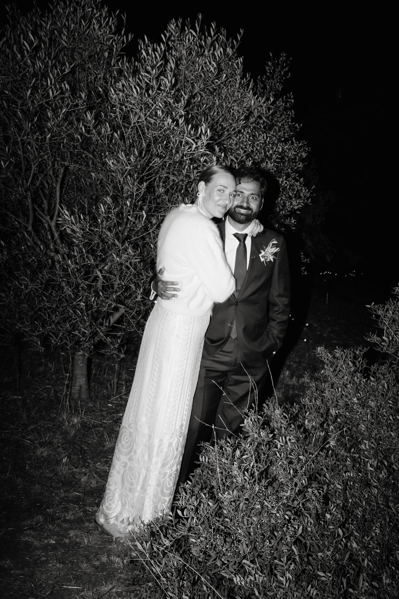 A black and white wedding photo of a couple embracing outdoors at night among garden foliage.