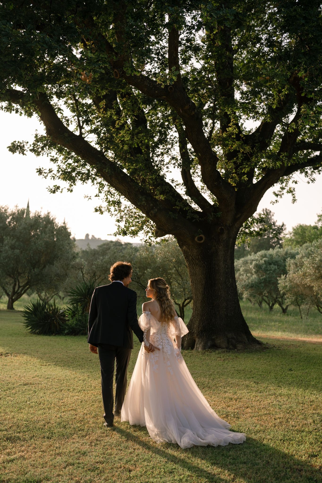 Couple walking hand in hand under a majestic old tree in a garden setting.