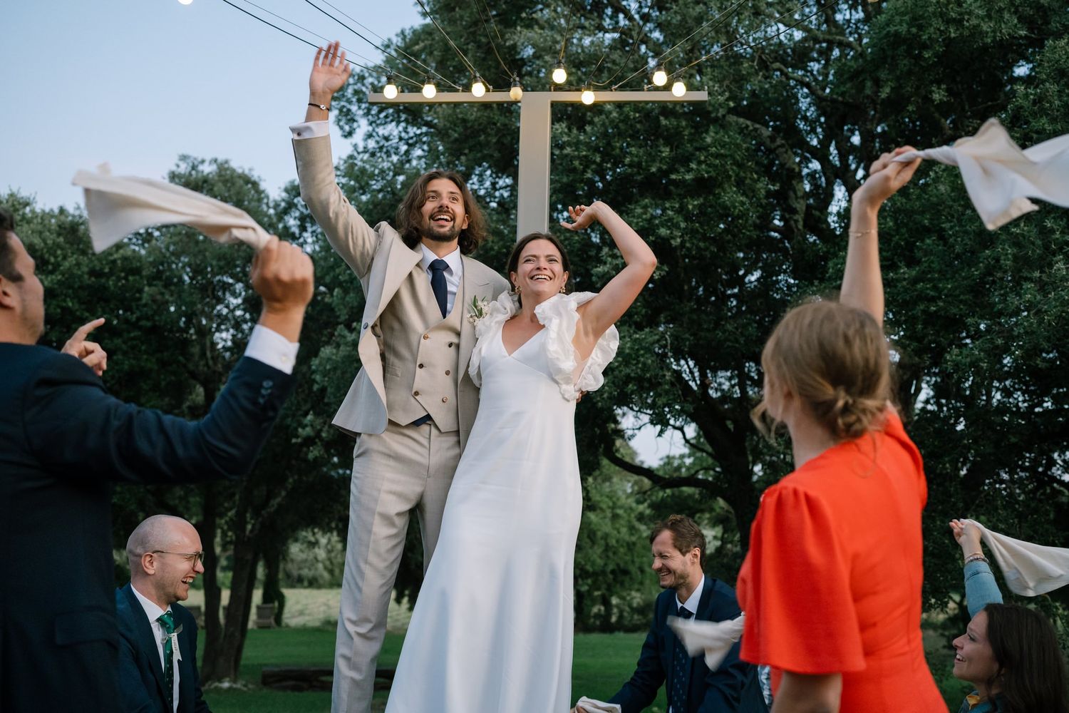 Wedding guests dance and celebrate outdoors under strung lights at dusk.