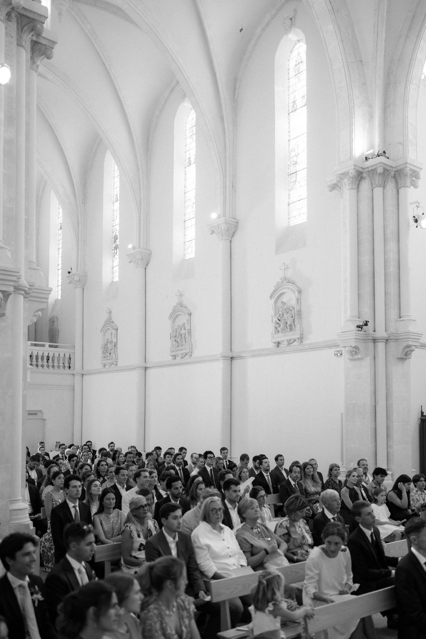Black and white photo of wedding guests filling the pews of a grand cathedral with tall windows.