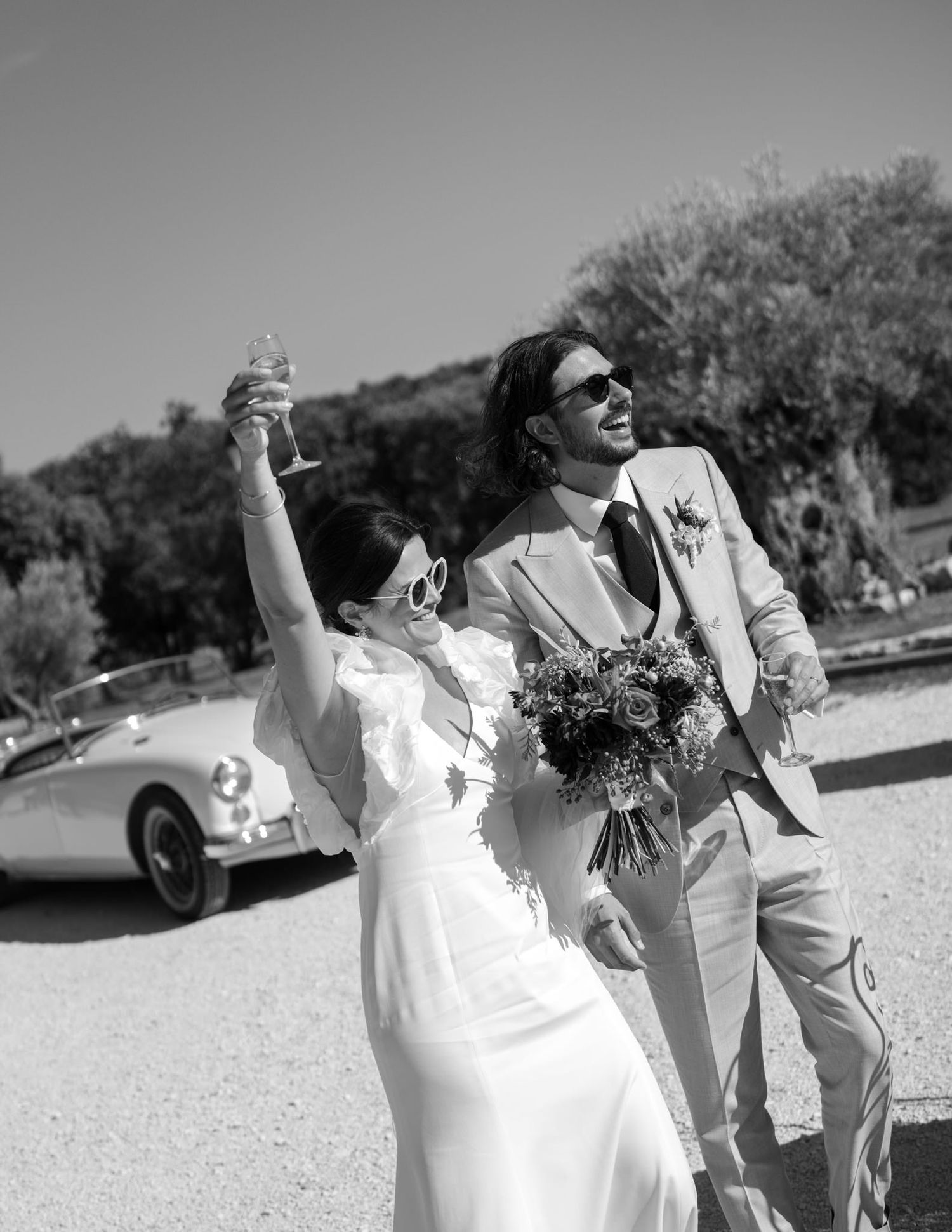 Black and white photo of joyful wedding celebration poses by classic car.