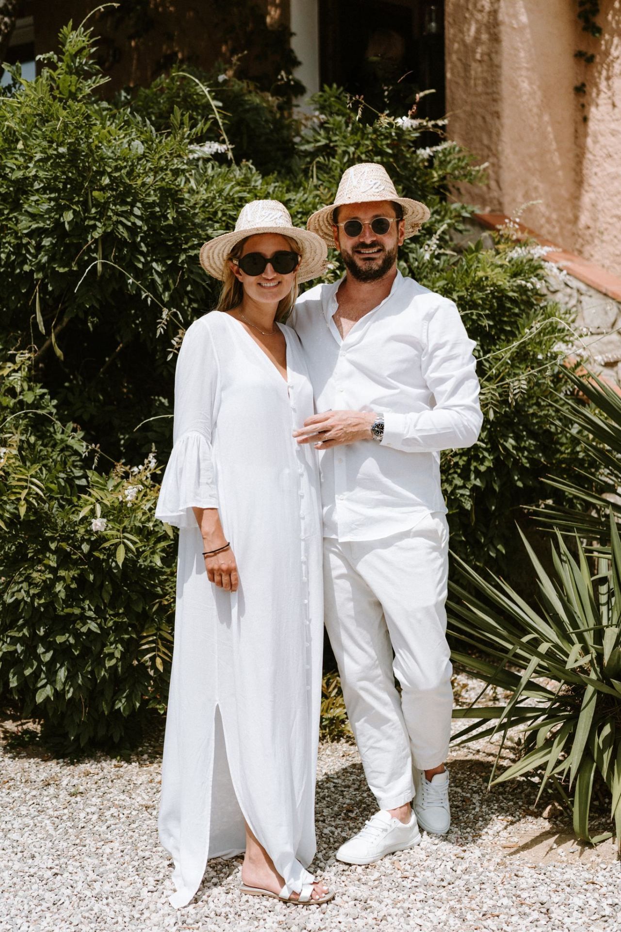 Couple in all-white outfits and sun hats posing together on a gravel path surrounded by greenery.