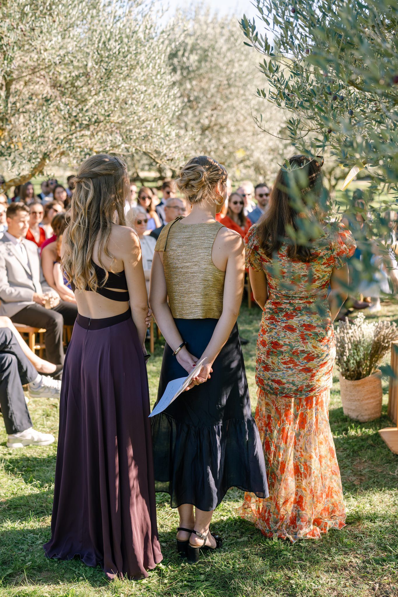 Wedding guests in colorful dresses mingle under olive trees during an outdoor celebration.