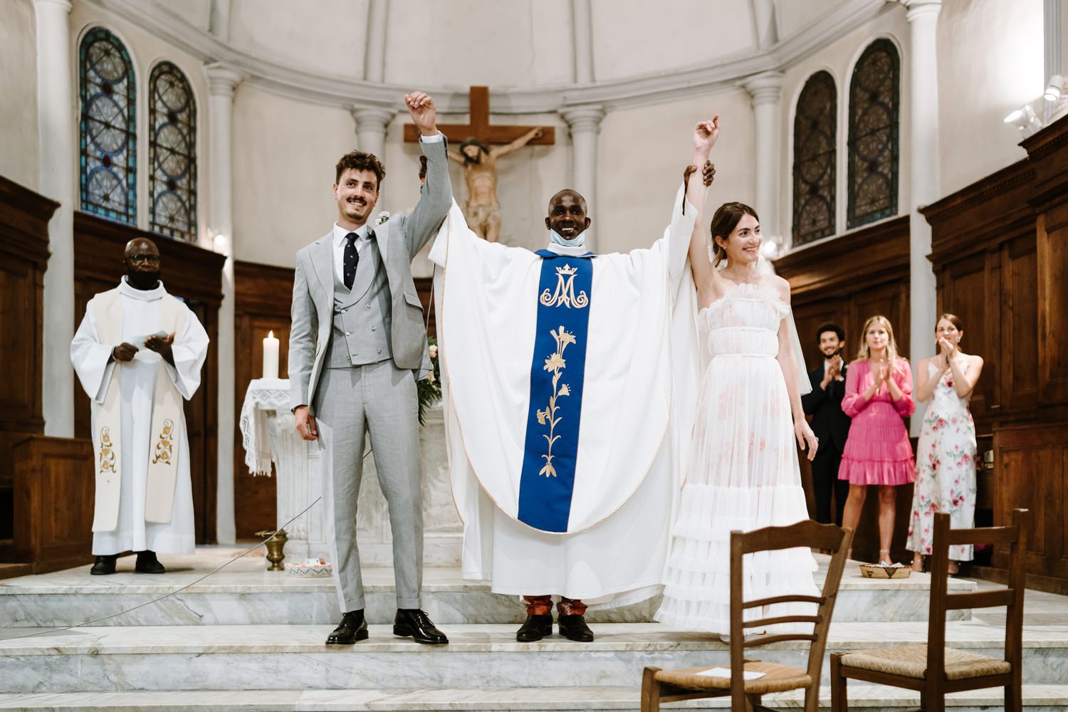 A celebratory moment at a church wedding with the couple and priest raising hands at the altar.