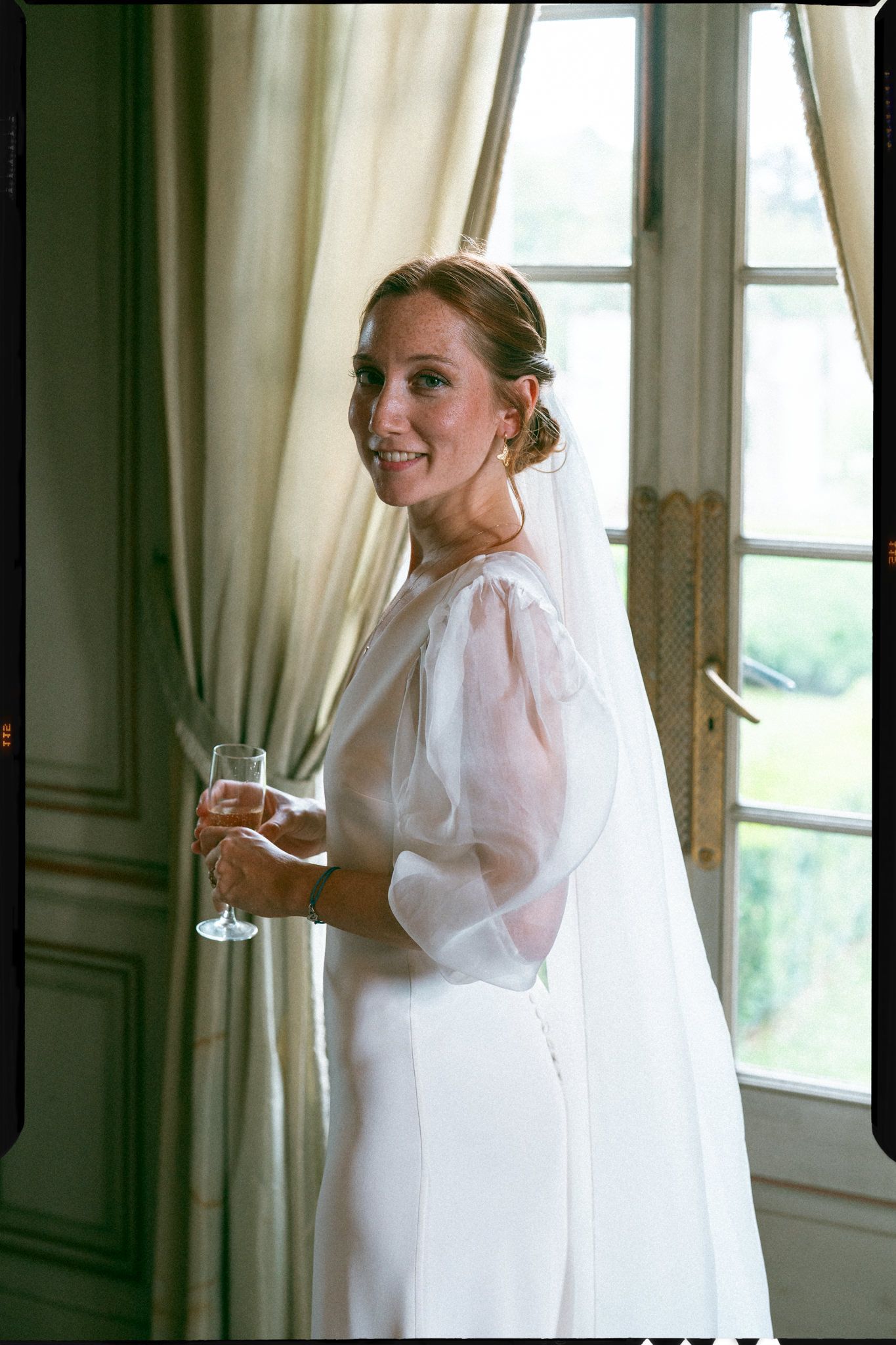 Bride in white dress with puffy sleeves poses by window with champagne glass in elegant room.