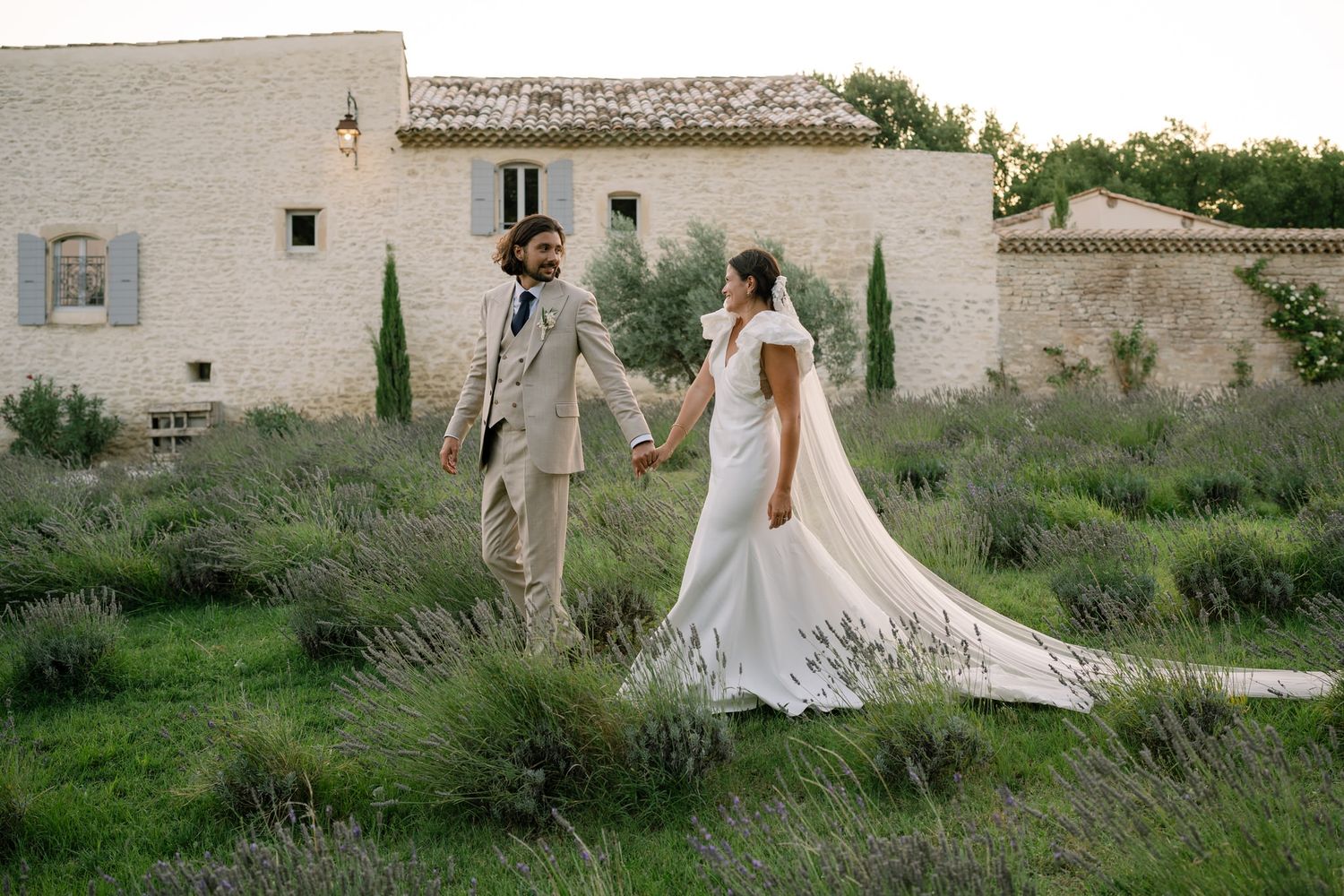 Newlyweds walk hand in hand through a rustic outdoor venue with stone buildings.