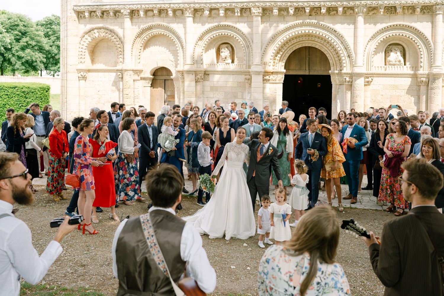 Crowd gathers outside ornate stone building for wedding celebration.