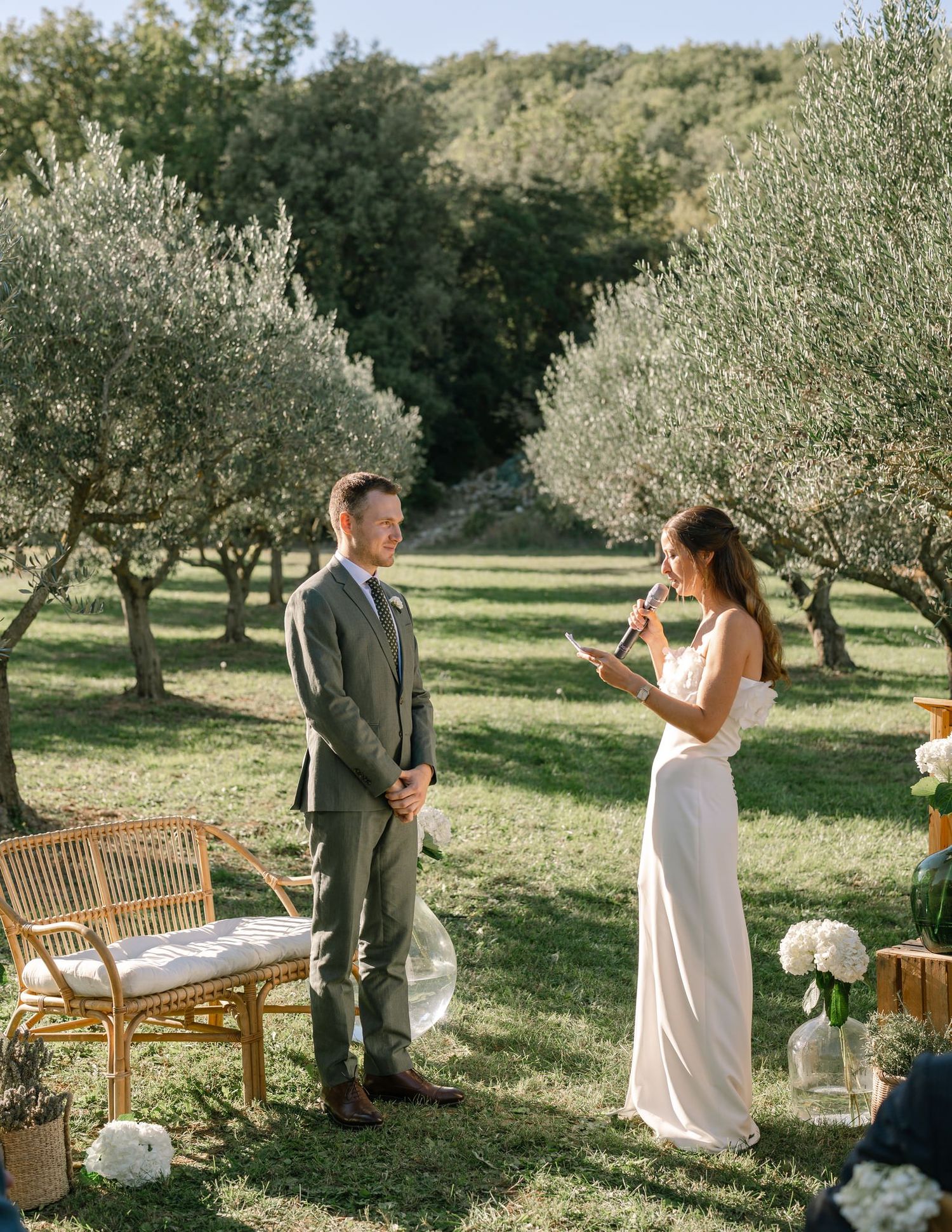 Intimate wedding ceremony taking place among olive trees with rustic wooden seating.
