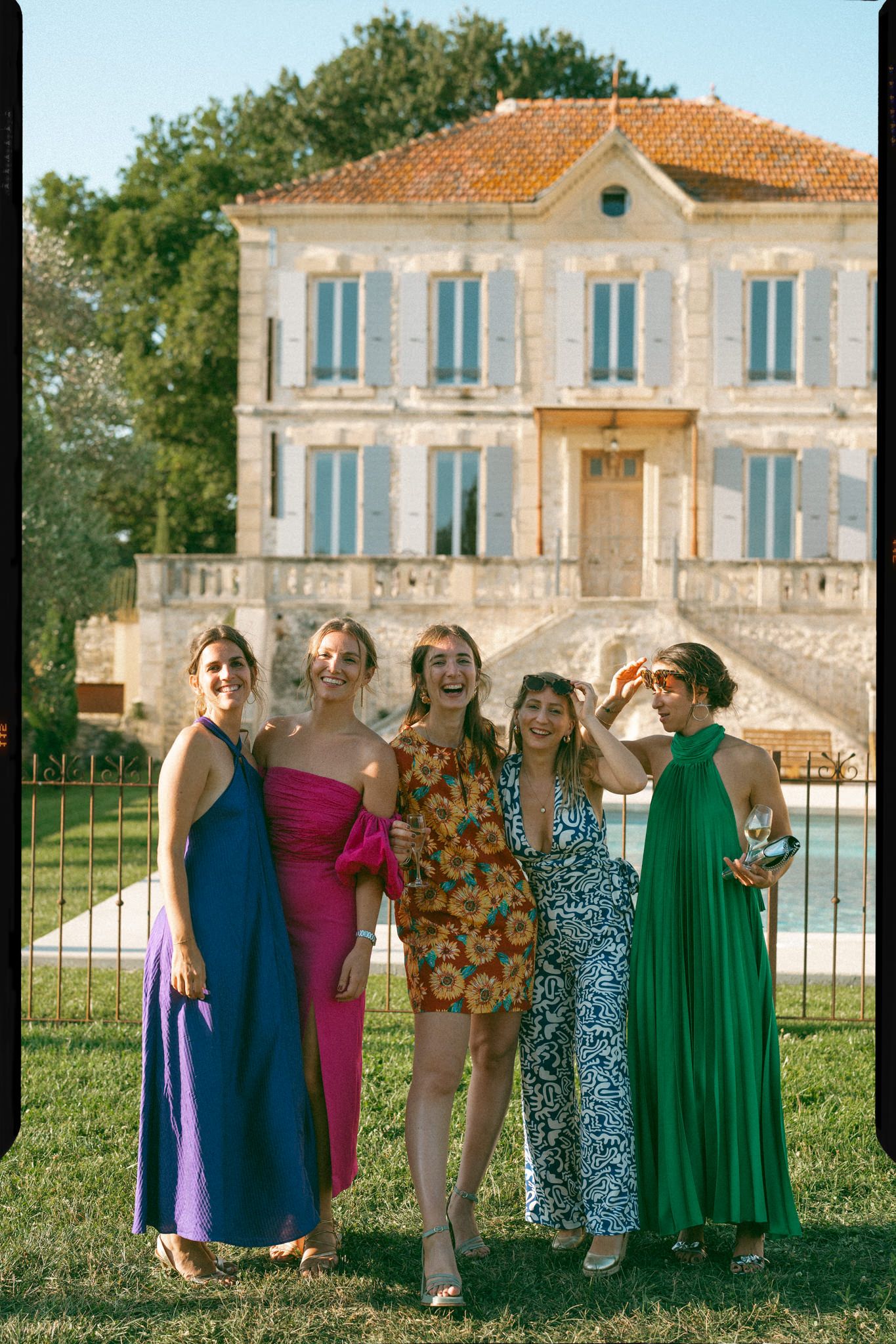 A group of people in colorful dresses and formal attire pose together in front of a Mediterranean-style villa.