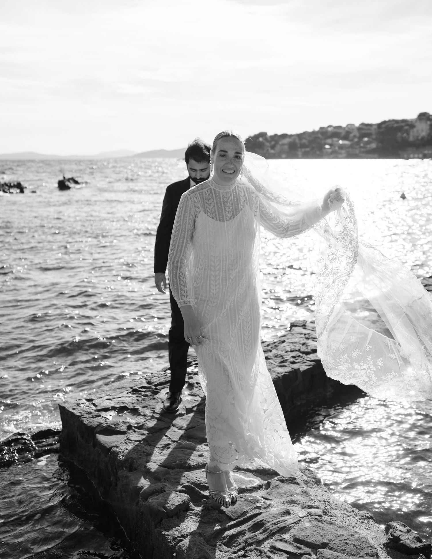 Two figures in white stand on coastal rocks with ocean waves splashing behind them.