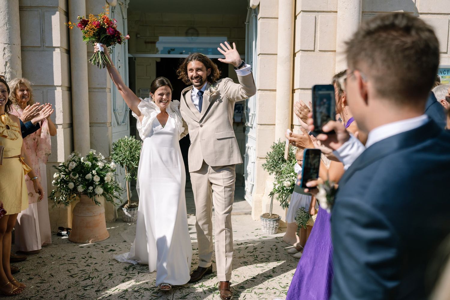 Newlyweds exit their wedding ceremony while guests celebrate with confetti at a beautiful stone building entrance.